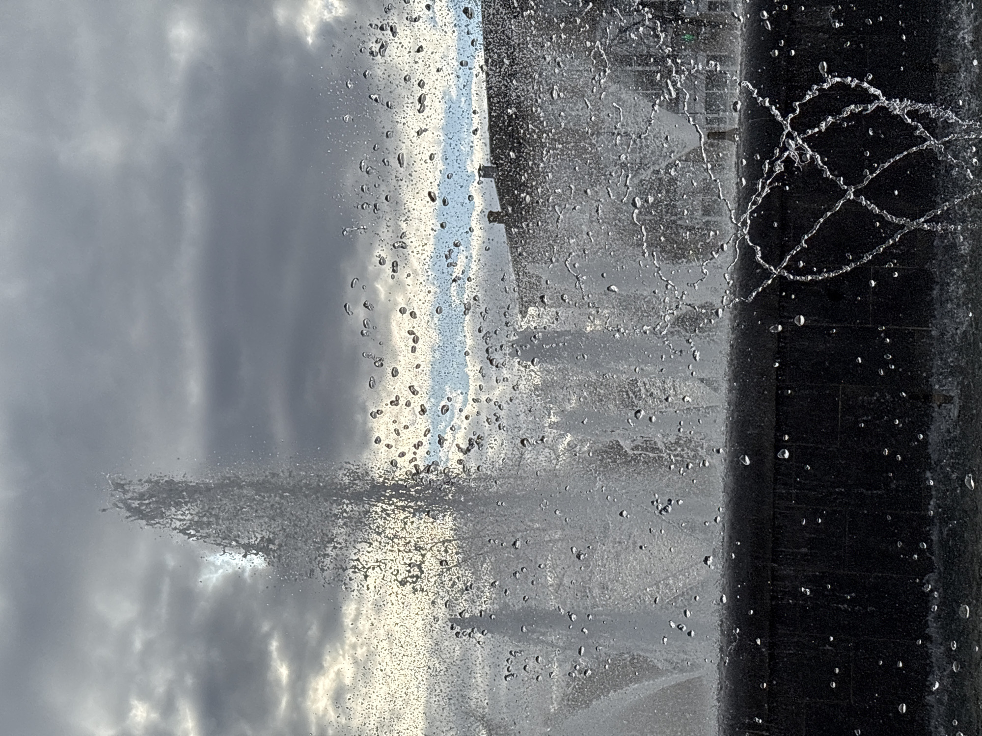 The image depicts a fountain with water droplets suspended in mid-air, partially obscuring a cloudy sky. A dark, vertical structure, likely part of the fountain, is visible on the right side. In the background, a hint of blue sky can be seen between the water spray. The image may be related to the city of Chaikovsky, Russia.