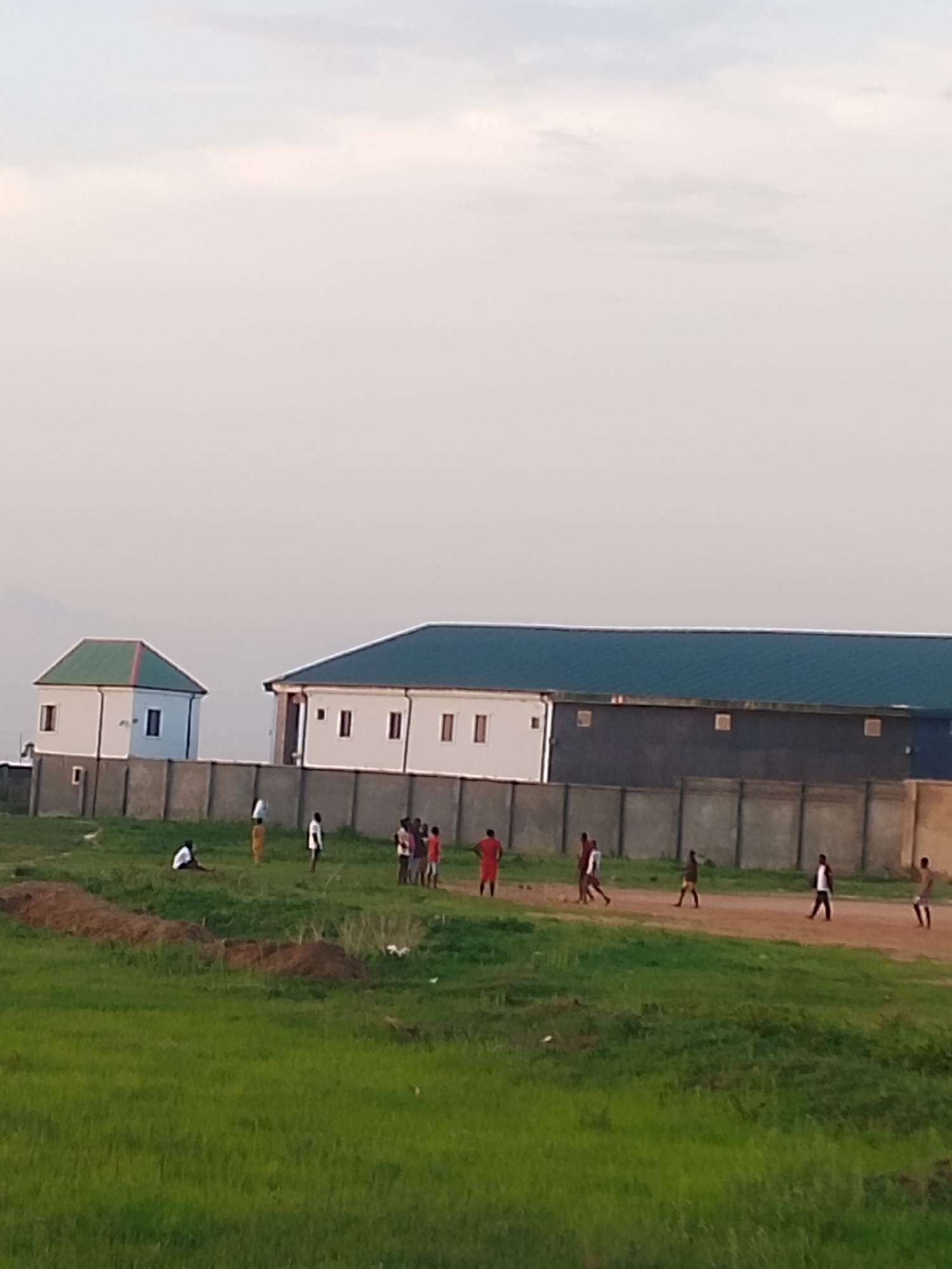 In Ibo, Nigeria, a group of approximately 12 people are present on a grassy field. Behind them are two buildings with green roofs, separated by a concrete wall. The sky is overcast. Some of the individuals appear to be walking or standing, while others are engaged in an unknown activity. The ground is primarily green grass, with patches of bare earth.
