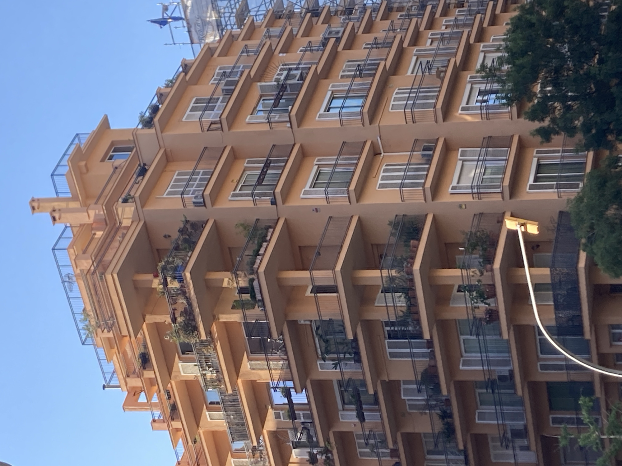 This is a slightly tilted, wide-angle, outdoor shot of a modern, peach-colored apartment building with many balconies. The building appears to be very tall and has a unique, stepped architectural design with staggered balconies. Many of the balconies have railings and some have plants, giving a sense of greenery and life on the exterior. Small air conditioning units are visible on some balconies, as are some windows and doors. Scaffolding is visible at the very top of the building, suggesting ongoing construction or renovation. To the right of the building, the branches of a large, dark green tree are visible, partially obscuring some of the upper floors. The sky is a clear, bright blue, indicating a sunny day. A light post is visible on the right side of the frame, with a curved white cable extending from it. The image gives a sense of urban living in a modern, somewhat abstract architectural setting. There is no visible text in the image.