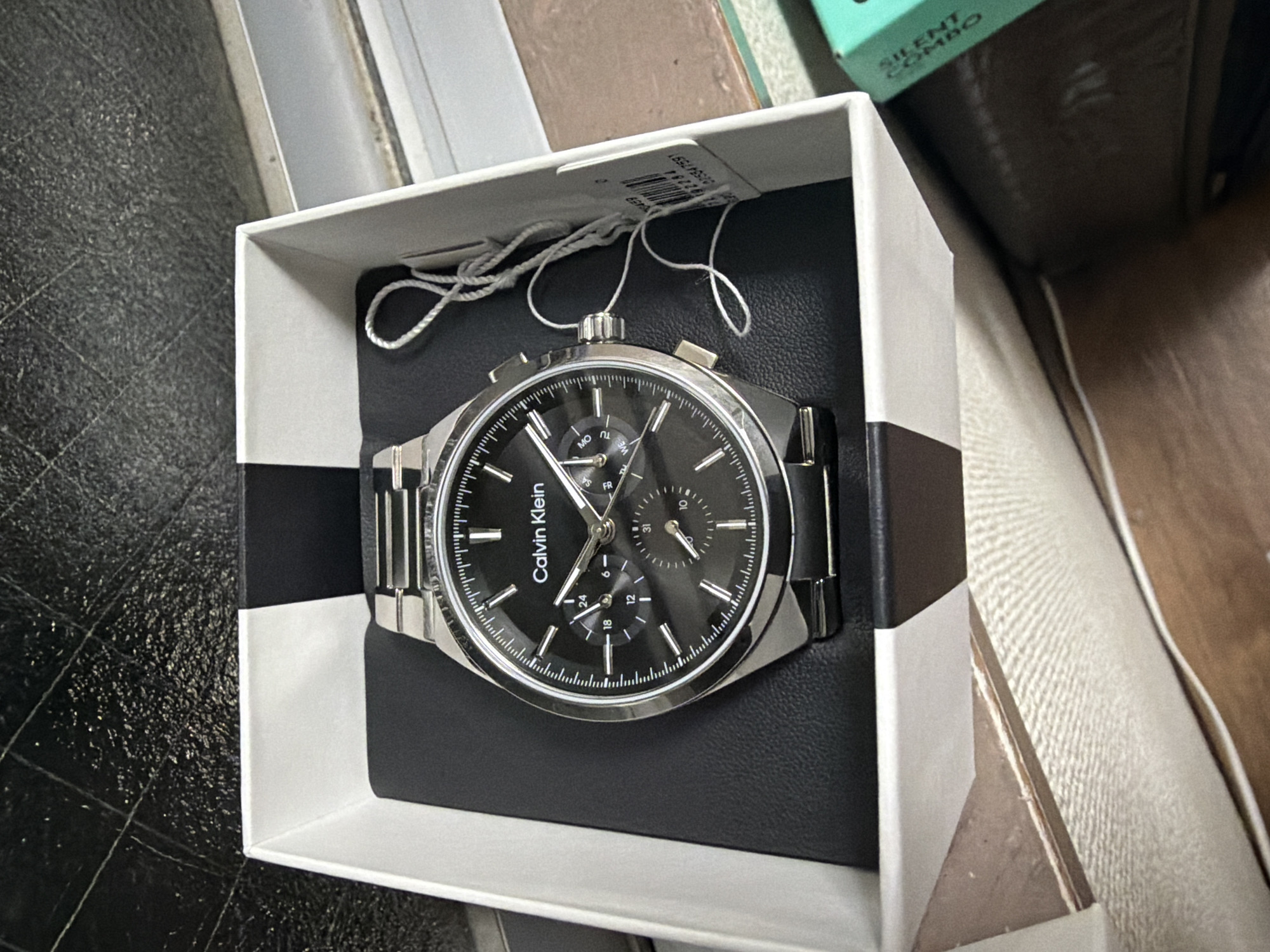 A Calvin Klein chronograph watch with a black dial and silver-tone metal bracelet is presented in its original white and black cardboard packaging. The watch is centrally positioned within the open box, which rests on a dark textured surface that appears to be the interior lining of the box. A white string tag, commonly attached to new watches, is loosely looped to the right of the watch. The background is a dark, textured material, possibly leather or a similar embossed surface, with faint grid lines visible. The packaging is placed near an opening that reveals a blurred view of the environment outside the box. The scene is observed from a slightly elevated, frontal perspective. No people are present in the image. The context of Adachi, Japan is not discernible from the visual information provided.