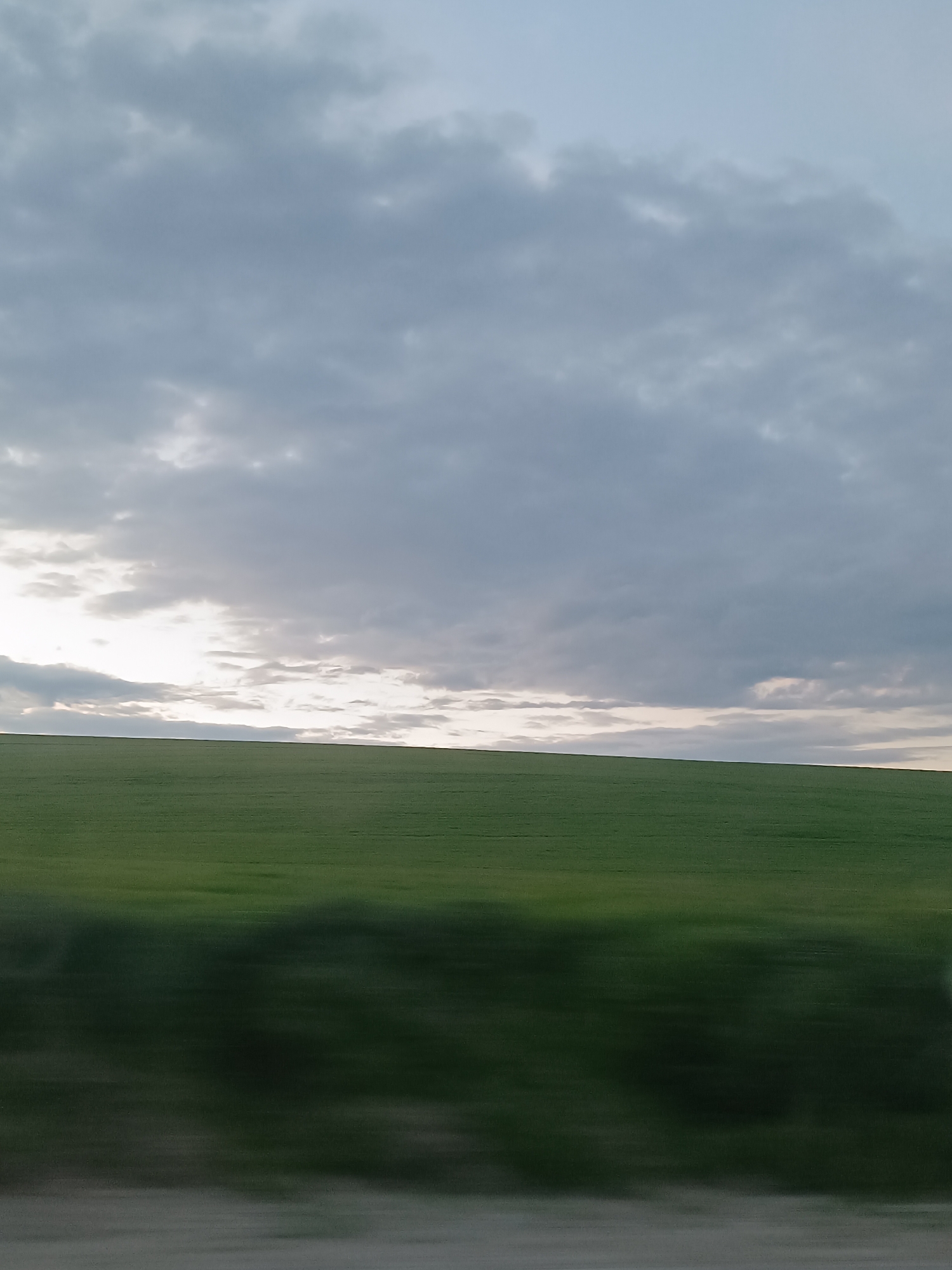 The image depicts a landscape under a cloudy sky. A green field dominates the scene, its lower portion blurred, suggesting movement or speed during image capture. The sky is filled with gray clouds, and a sliver of light is visible near the horizon line. The photo appears to be taken from a moving vehicle, likely in a rural area. The location is identified as Unknown, Unknown, which means there is not enough information to determine where the photo was taken.