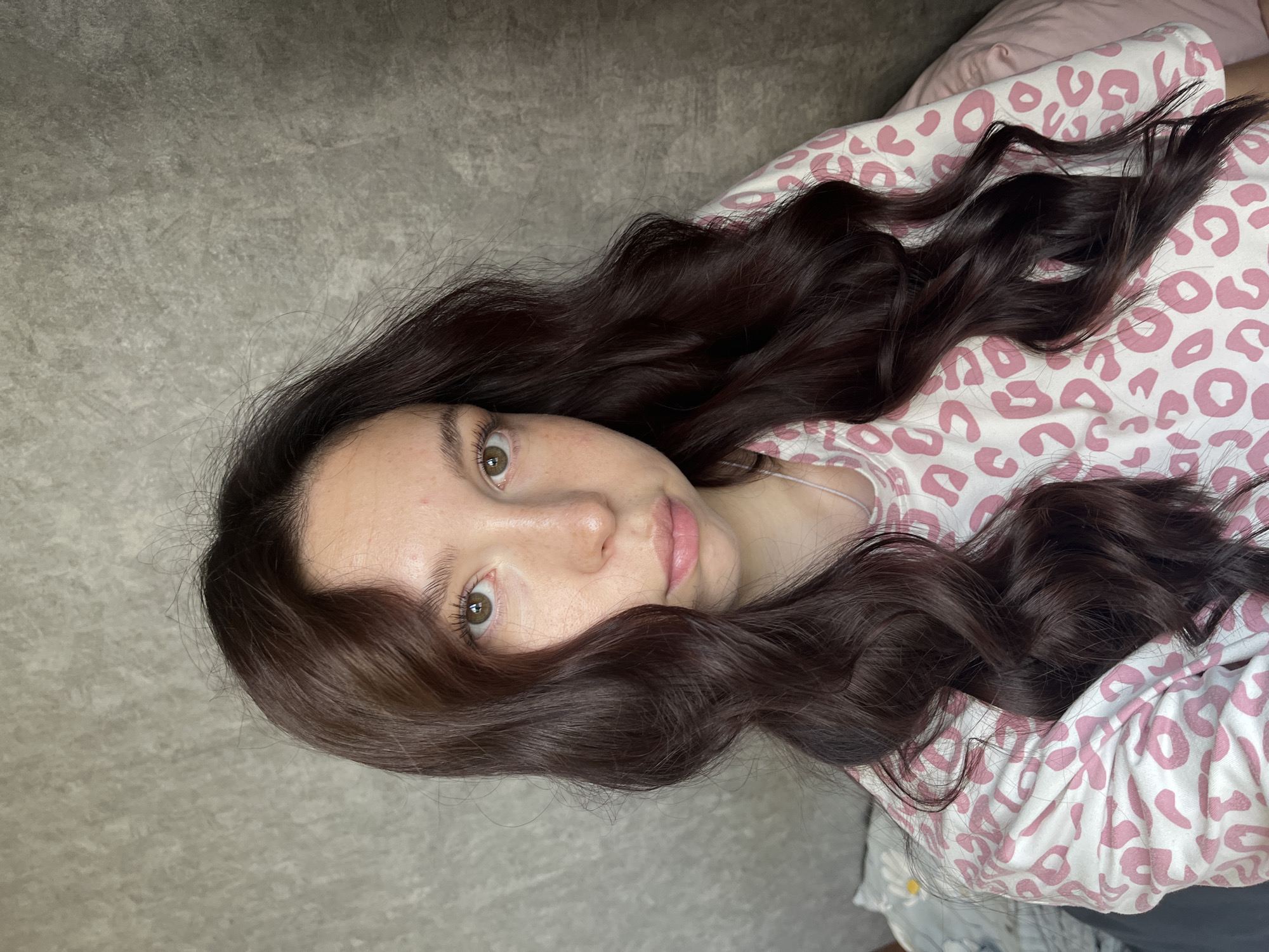 The image is a close-up, head-and-shoulders shot of a young woman with dark, wavy hair and light skin with freckles. She is wearing a white long-sleeved shirt with a pink leopard print pattern. The background is a textured grey wall. The woman is looking directly at the camera with a neutral expression. The lighting is soft and diffused, suggesting an indoor setting. There are no specific elements in the image that indicate a location in Chaikovsky, Russia.