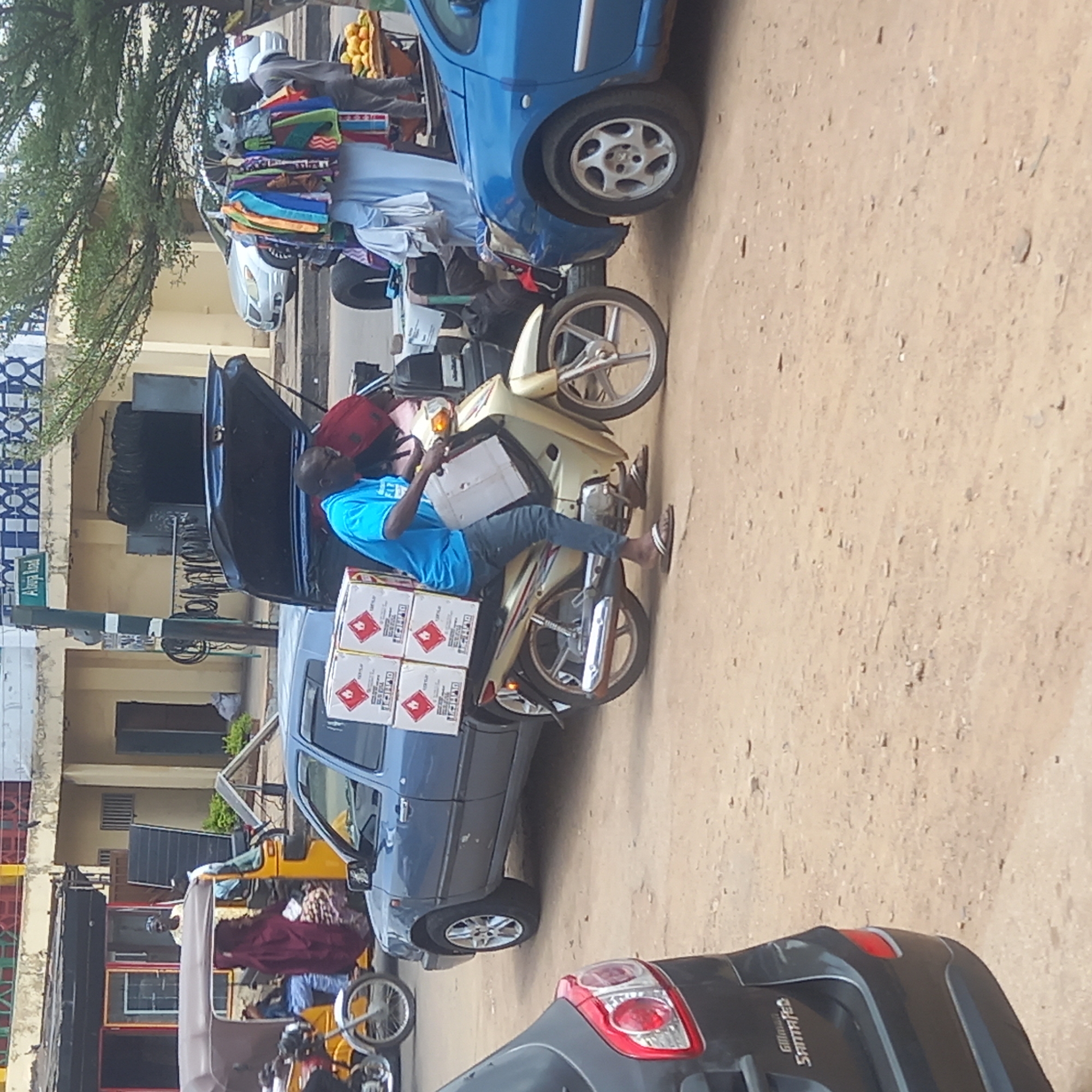 In Dadi Riba, Nigeria, a man wearing a blue shirt and sandals sits on a beige motorcycle. He is holding a box. Behind him are four additional boxes stacked on the motorcycle. The motorcycle is parked between a dark grey car with its trunk open and a light blue car. A yellow tricycle taxi is visible to the left of the motorcycle. On the far left is a sign with the words "Abuja Road". The ground is unpaved and appears to be a dirt road.