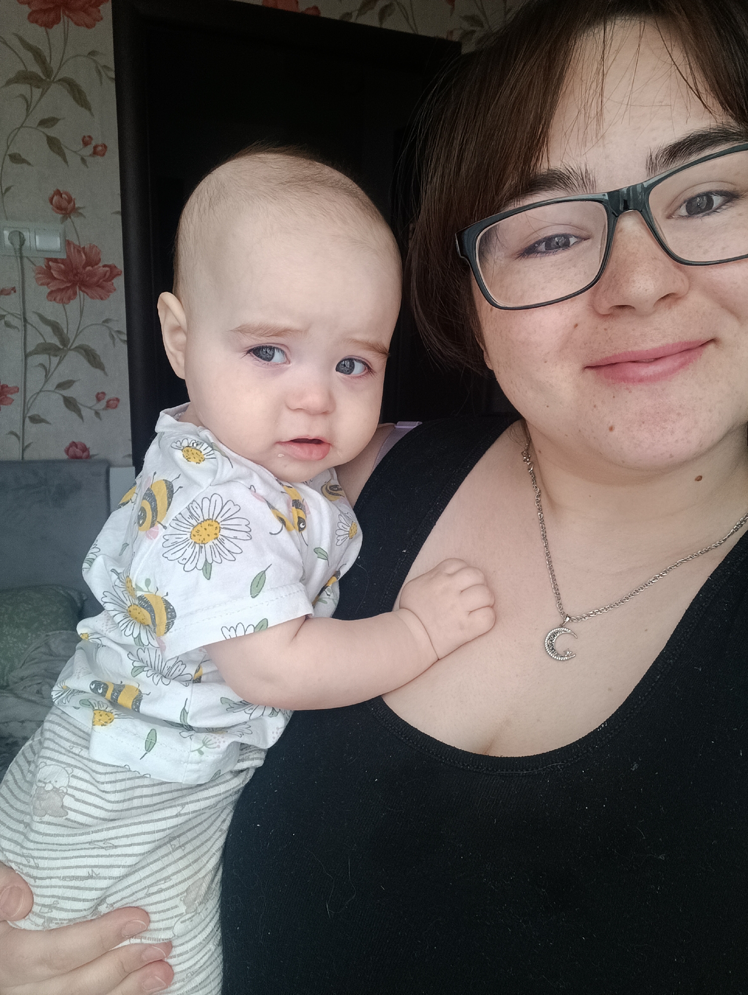 A waist-up, close-up shot captures a woman holding a baby. The woman is fair-skinned with freckles, short brown hair, and glasses. She is wearing a black top and a silver necklace with a crescent moon pendant. She smiles faintly at the camera.
The baby has fair skin, light eyebrows, and blue eyes. The baby wears a white shirt adorned with daisies and bees, and striped pants. The baby looks directly at the camera with a slightly worried expression.
The background features wallpaper with a floral pattern in muted red and green tones. A dark doorframe is visible behind the baby's head. To the left, a gray headboard and part of a pillow are visible. The exact location and city cannot be determined.