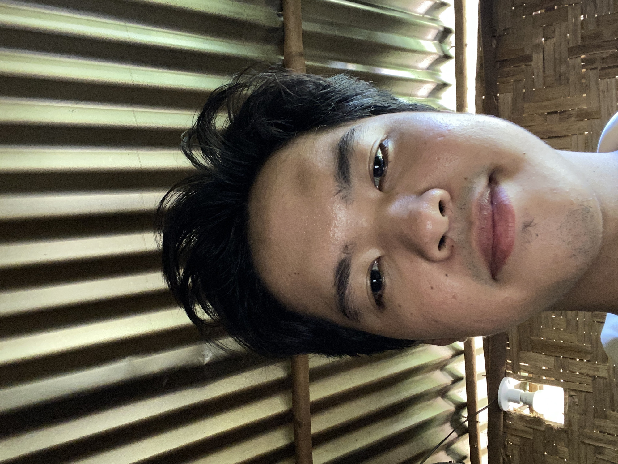 A close-up, upward-facing selfie of a young man's face. He has dark hair and fair skin, with visible pores and a few small blemishes on his forehead and cheeks. His eyes are dark and looking directly at the camera. The man's lips are slightly parted. The background consists of a corrugated metal roof with distinct vertical ridges, casting striped shadows. Below the roof, a woven bamboo wall is visible, with a single lightbulb hanging from a wire. The scene appears to be outdoors or in a structure with open sides, possibly in a tropical environment, suggesting a location within Davao City, Philippines.