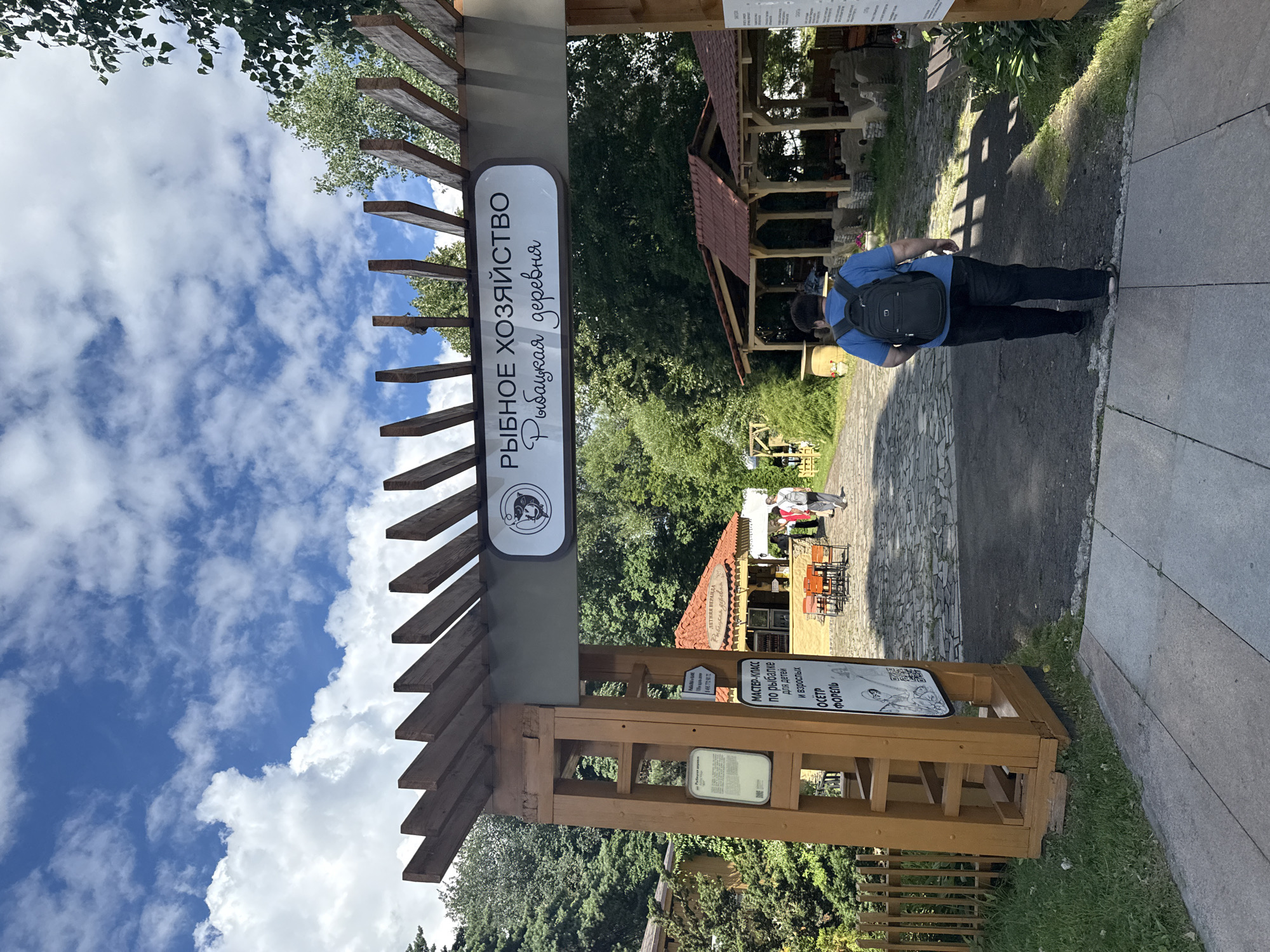 The image shows an entrance archway to a "Fishing Farm - Fisherman's Village" in Moscow, Russia. The archway is constructed of wooden beams and has a sign overhead with Russian text and a logo of a fish. To the left of the entrance is a wooden post with several signs, one of which advertises a master class in fishing for children and adults. In the background, there are covered outdoor seating areas with tables and chairs, and lush green trees and vegetation. A person wearing a blue shirt and black pants, carrying a black backpack, is walking away from the camera through the archway. The sky is blue with scattered white clouds.
