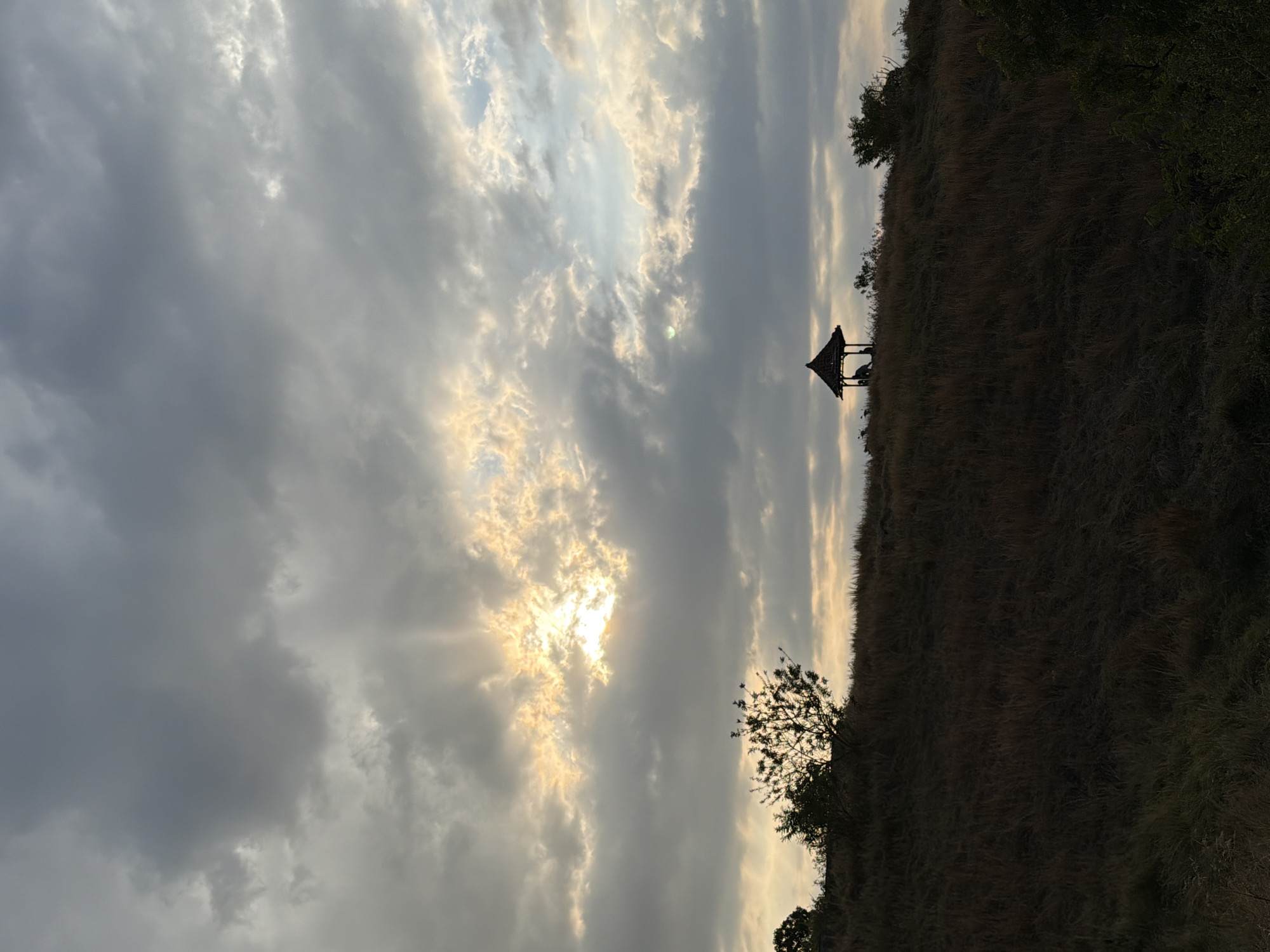 The image depicts a landscape with a silhouetted gazebo on a grassy hill under a cloudy sky. The sun is partially visible through the clouds, casting rays of light. There are trees on the hill, and the foreground is filled with dry grass. The scene appears to be in a rural or natural setting. The presence of a gazebo suggests a potential viewpoint or resting spot. The overall mood is atmospheric and serene, indicative of either sunrise or sunset. The location is identified as Kubutambahan, Indonesia.