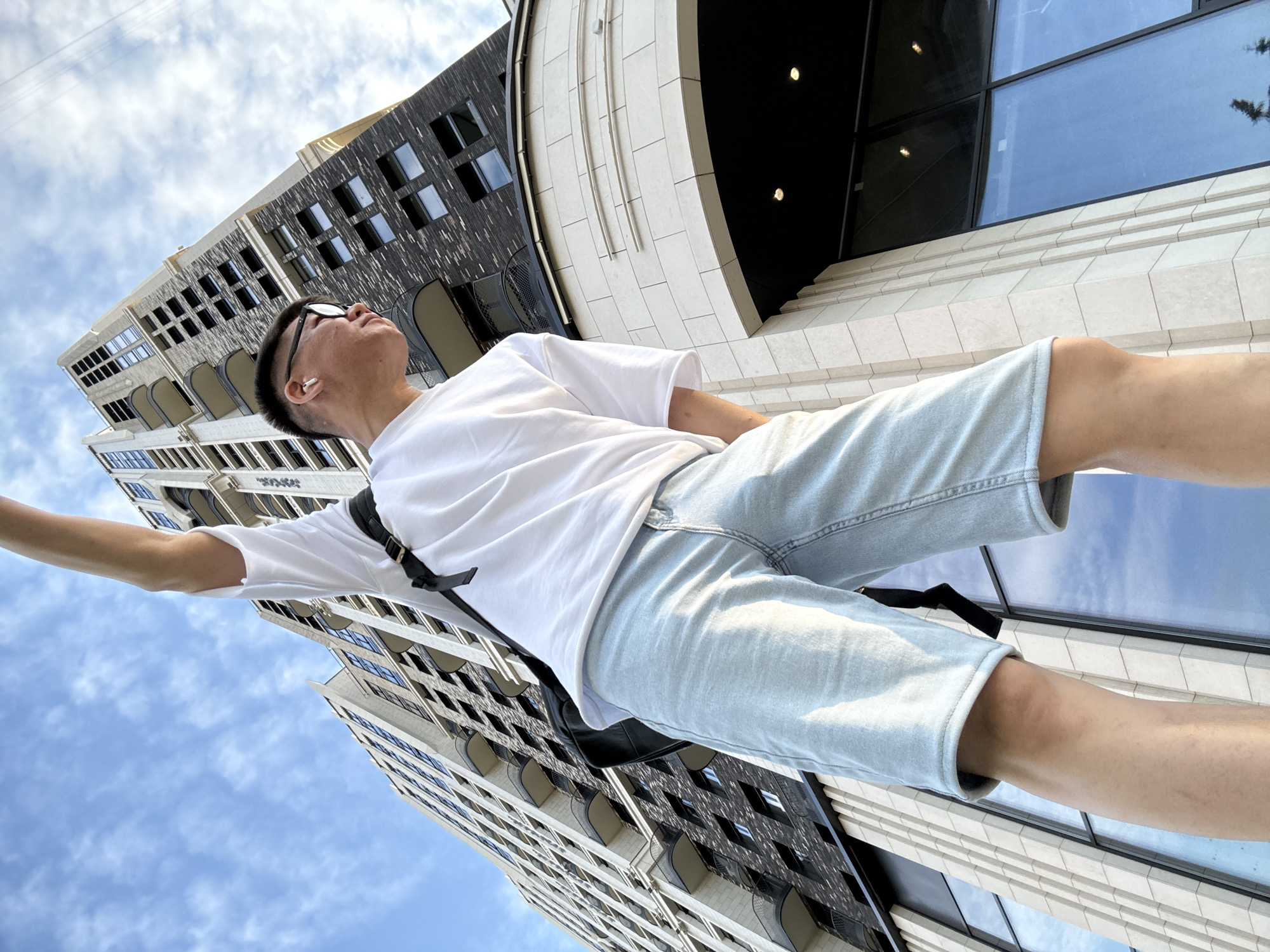 A young Asian man stands in front of a modern building, his arm raised towards the sky. He is wearing a white t-shirt, light blue denim shorts, and glasses, with white earbuds in his ears. A black strap, likely from a backpack, is visible across his back. The building appears to be a tall residential complex, with a mix of light and dark facade materials, featuring numerous windows and balconies. The sky above is blue with scattered clouds. The perspective of the shot is from below, looking up at the man and the building, emphasizing their scale. The image was captured on a sunny day, with soft shadows indicating the direction of the light. The architecture suggests a contemporary urban setting.