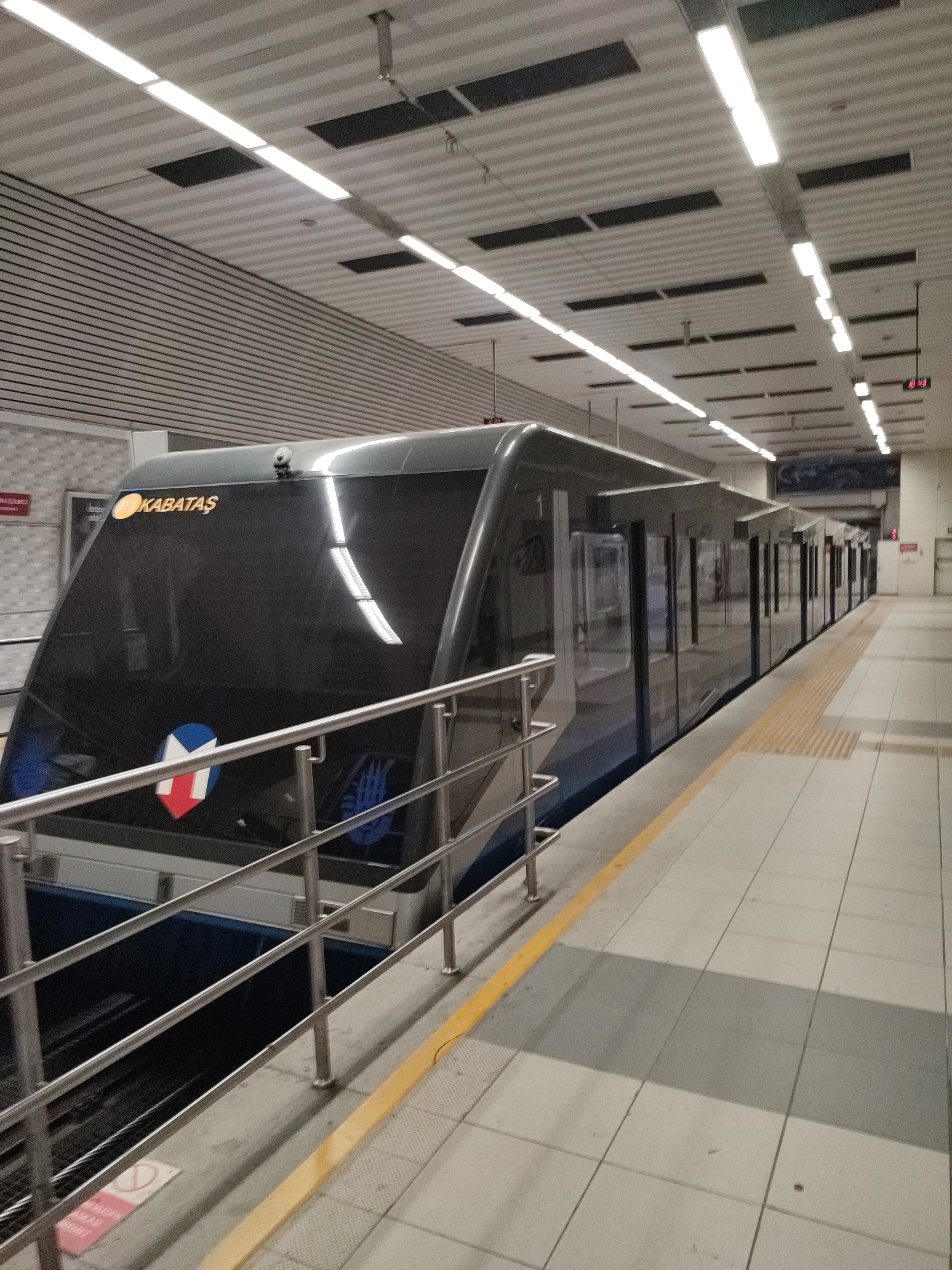 The image captures an interior view of a modern metro station in Beyoğlu, Turkey, specifically the Kabataş funicular station. A sleek, dark gray funicular train is parked at the platform, its large, tinted windows reflecting the station's bright fluorescent lighting. The train is oriented diagonally across the frame, from the lower left to the upper right. The front of the train displays an illuminated "KABATAŞ" sign and a circular emblem featuring red, white, and blue geometric shapes. A metal railing runs along the edge of the platform in front of the train. The platform itself is paved with light-colored square tiles, with a yellow tactile warning strip running along the edge. Above, the ceiling features a ribbed metal design with numerous rectangular fluorescent light fixtures and dark rectangular ventilation grilles. In the background, a digital clock displays "10:43." The overall environment appears clean, well-lit, and contemporary. There are no people visible in the frame.