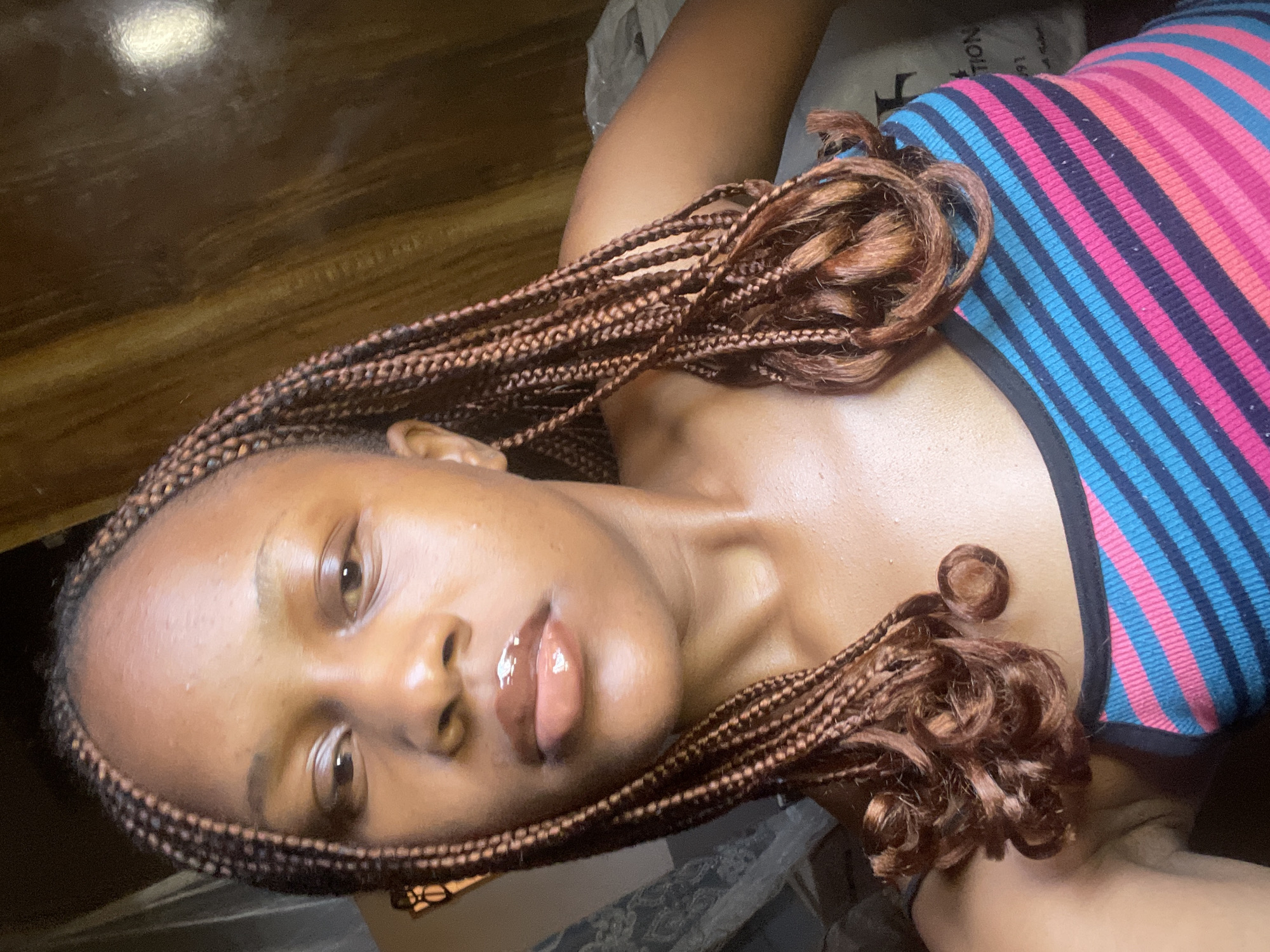 A young woman with braided hair is pictured in a close-up shot. She is looking directly at the camera, her expression neutral. Her lips are glossy and her skin appears to be dewy. She is wearing a multi-colored striped tank top. The background is dimly lit and appears to be an indoor setting, possibly a room with curtains and some stored items. The image's quality suggests it was taken with a mobile phone. Without additional context or visual cues, it is not possible to determine the specific location within Jos, Nigeria.