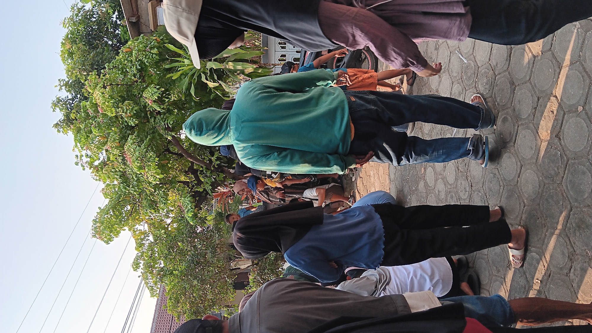The image depicts a group of people in an outdoor setting, likely a residential area in Cerme, Indonesia. The ground is paved with hexagonal gray stones. In the foreground, several individuals are visible from behind. On the left, a person wearing a dark gray long-sleeved shirt stands with their back to the camera. To their right, a person in a blue long-sleeved shirt and black pants, wearing a black hijab, stands with their arms crossed. In the center, a person in a teal hooded sweatshirt and blue jeans faces away from the viewer. To the right, a person wearing a beige baseball cap and a dark hijab is partially visible.
In the background, more people are gathered, including children. Some are standing under the shade of a large tree with green leaves and pink blossoms. To the left, a building with a tiled roof is visible. A portion of a motorcycle with a red rim is also present on the right side of the image. The overall atmosphere appears to be informal, suggesting a community gathering or event. The lighting suggests it is daytime, likely late afternoon due to the warm tones and shadows.