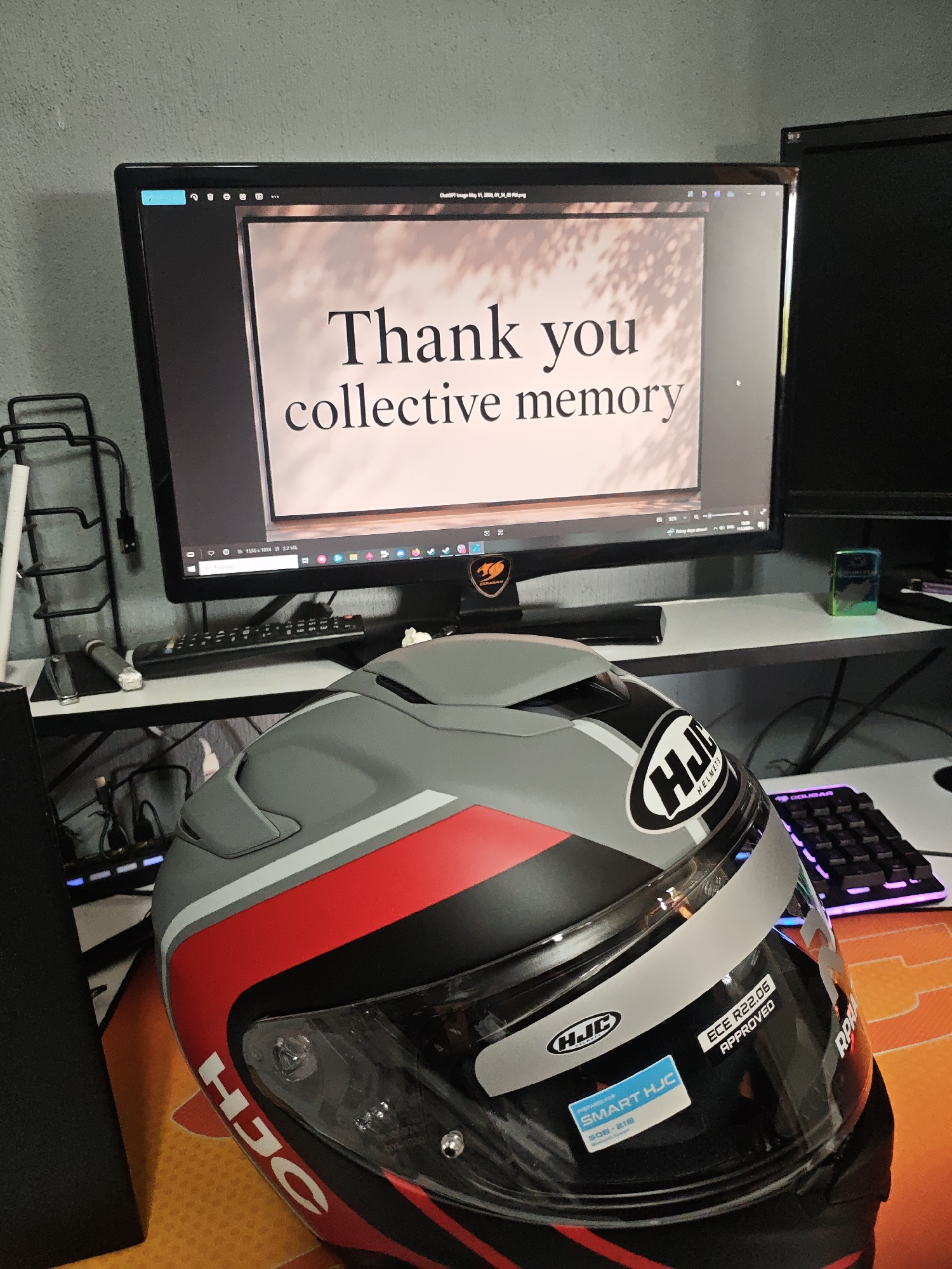 The image shows a desk setup with a computer monitor displaying the words "Thank you collective memory." In the foreground is a gray, red, and black HJC motorcycle helmet. The HJC logo is visible on the side of the helmet along with stickers that say "SMART HJC" and "ECE R22.06 APPROVED." A black keyboard with illuminated keys is partially visible on the right side of the desk. Other items visible in the background include a second monitor, a wire rack, and various cables. The location in the city is Unknown, Unknown.