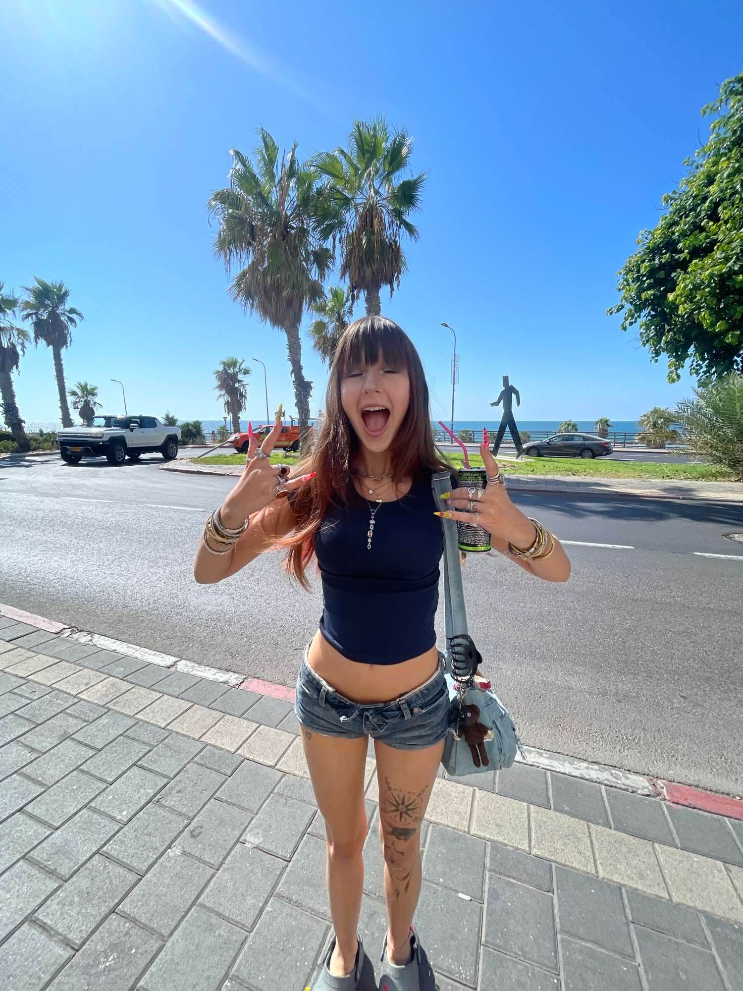 A young woman with long brown hair and bangs stands on a paved sidewalk, facing the camera. She is wearing a dark blue cropped tank top, denim shorts, and dark-colored slides. She has several rings on her fingers and bracelets on her wrists. Her right hand is raised, holding a can with a straw and making a peace sign. Her mouth is open, and she appears to be speaking or laughing. A light blue bag with a small brown bear charm is slung over her shoulder. Behind her, a street runs parallel to the sidewalk, with palm trees and a few vehicles. In the distance, the sea is visible. The scene appears to be in Tel Aviv, Israel, suggested by the architecture and vegetation typical of the city.