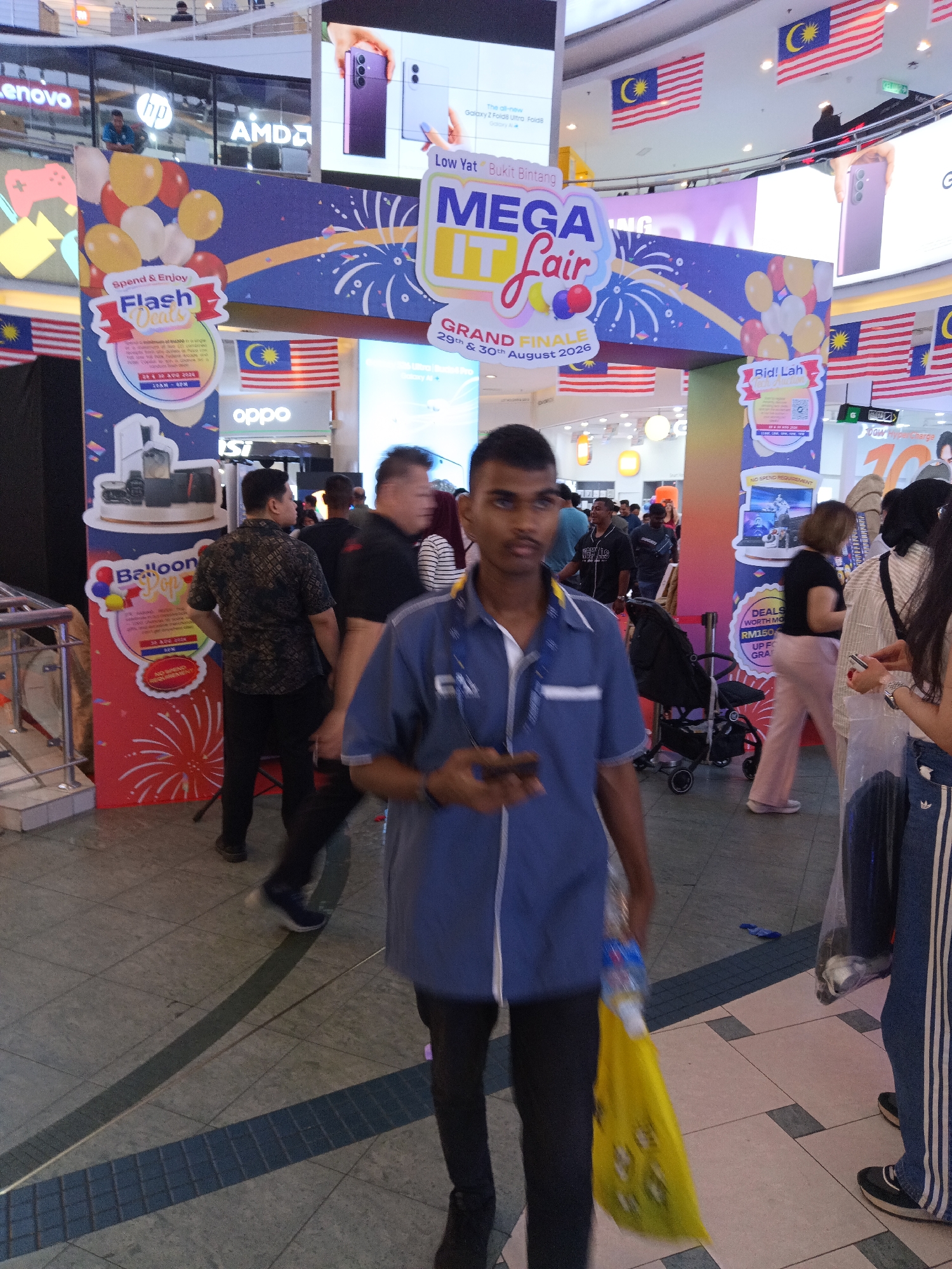 The image depicts the interior of a shopping mall, likely in Kuala Lumpur, Malaysia, with a vibrant "Mega IT Fair" in progress. Malaysian flags are prominently displayed throughout the venue, indicating its location. In the foreground, a young man in a blue uniform is walking towards the camera, holding a yellow plastic bag and a mobile phone. He appears to be an employee or attendee of the fair. In the background, various stalls and displays are visible, promoting electronics and technology products. People are browsing and shopping, suggesting a busy and engaging event. The overall atmosphere is lively and commercial, characteristic of a typical shopping mall event.