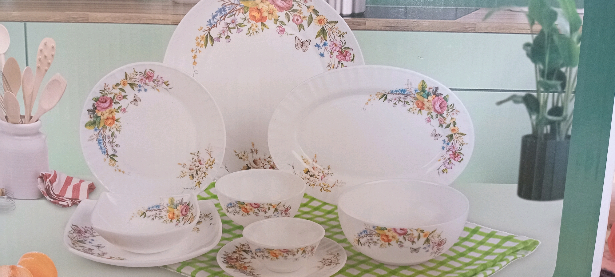 The image shows a white ceramic dinnerware set with a floral pattern. The set includes several plates of different sizes, bowls, and cups. The floral pattern features roses, daisies, and other small flowers in various colors, along with butterflies. The dinnerware is arranged on a light green and white checkered cloth. In the background, there is a hint of a light green wall and a plant in a dark pot. There are also some wooden kitchen utensils in a white ceramic holder on the left side of the image. The overall scene appears to be a product display or advertisement. There are no people visible in the image, and no specific location in Altynkul, Uzbekistan can be identified.