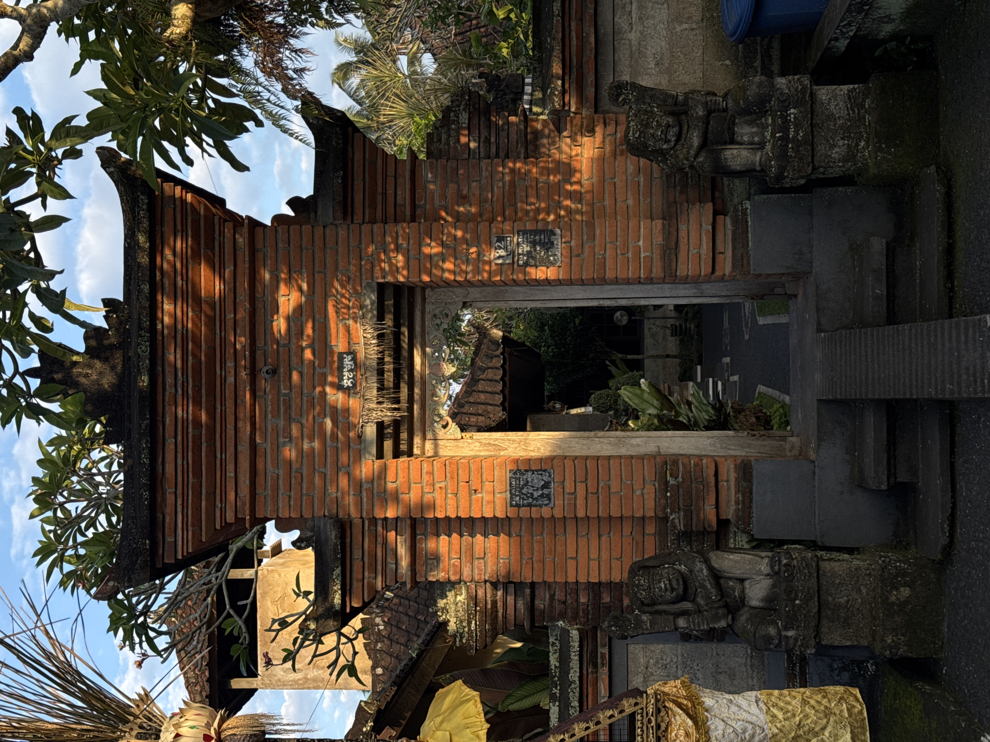 A traditional Balinese split gate made of red brick stands in Ubud District, Indonesia. The gate is flanked by two stone statues, one on each side, depicting guardian figures. The left statue is partially obscured by a decorative cloth. Sunlight casts shadows across the brickwork, highlighting the texture and details of the gate. Through the opening of the gate, a pathway leads into a garden with lush green foliage and a glimpse of a tiled roof. Above the gate, tropical trees with green leaves are visible against a blue sky with scattered clouds. A tall, light-colored building with a tiled roof is seen behind the left side of the gate, and a decorative yellow umbrella is visible near the left statue.
