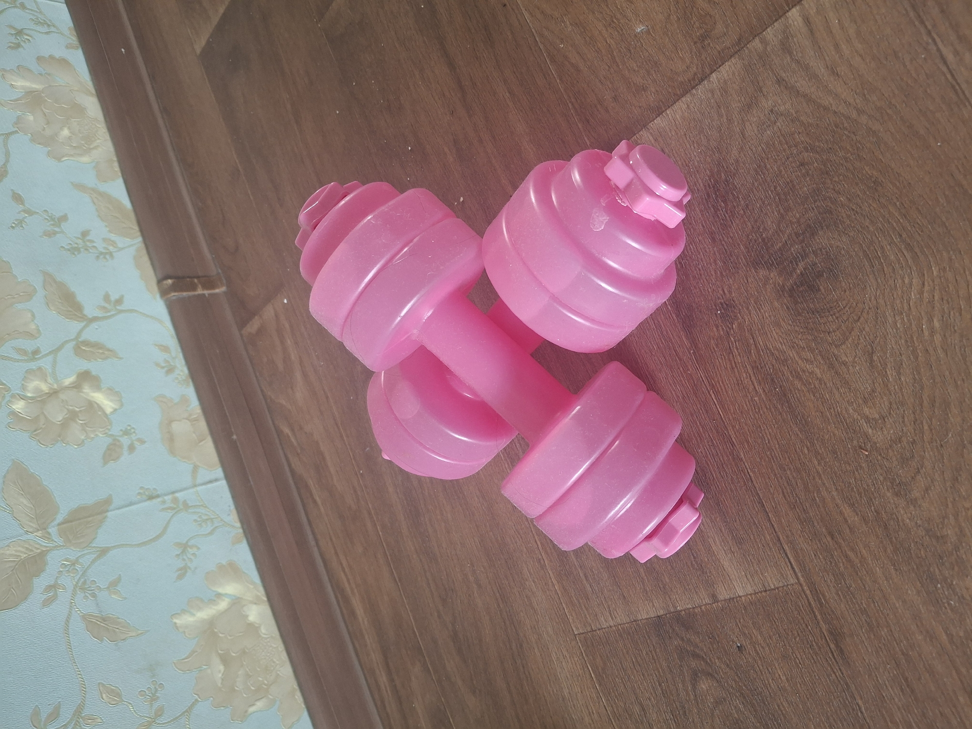 The image shows two pink dumbbells on a wooden floor. The dumbbells are plastic and have a ribbed design on the weight plates. In the background, there is a wall with floral wallpaper and a wooden baseboard. The scene appears to be indoors, likely in a residential setting. There is no indication of the location being in Aktobe, Kazakhstan.