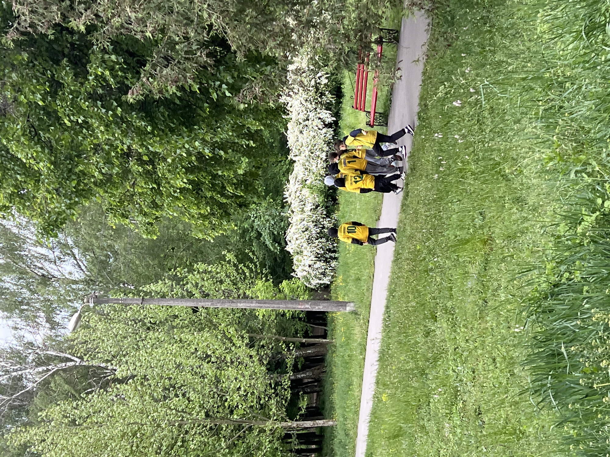A ground-level shot depicts a group of young individuals wearing yellow and black athletic outfits on a paved pathway. They are positioned closely together, possibly walking or jogging. The pathway is bordered on one side by lush green vegetation and on the other by trees and flowering bushes. A park bench with a red frame is also visible. A lamppost stands off to the side of the path. The location is within a green space, plausibly a park or recreation area. The image appears to be taken during daylight hours. The location is Ternopil, Ukraine.