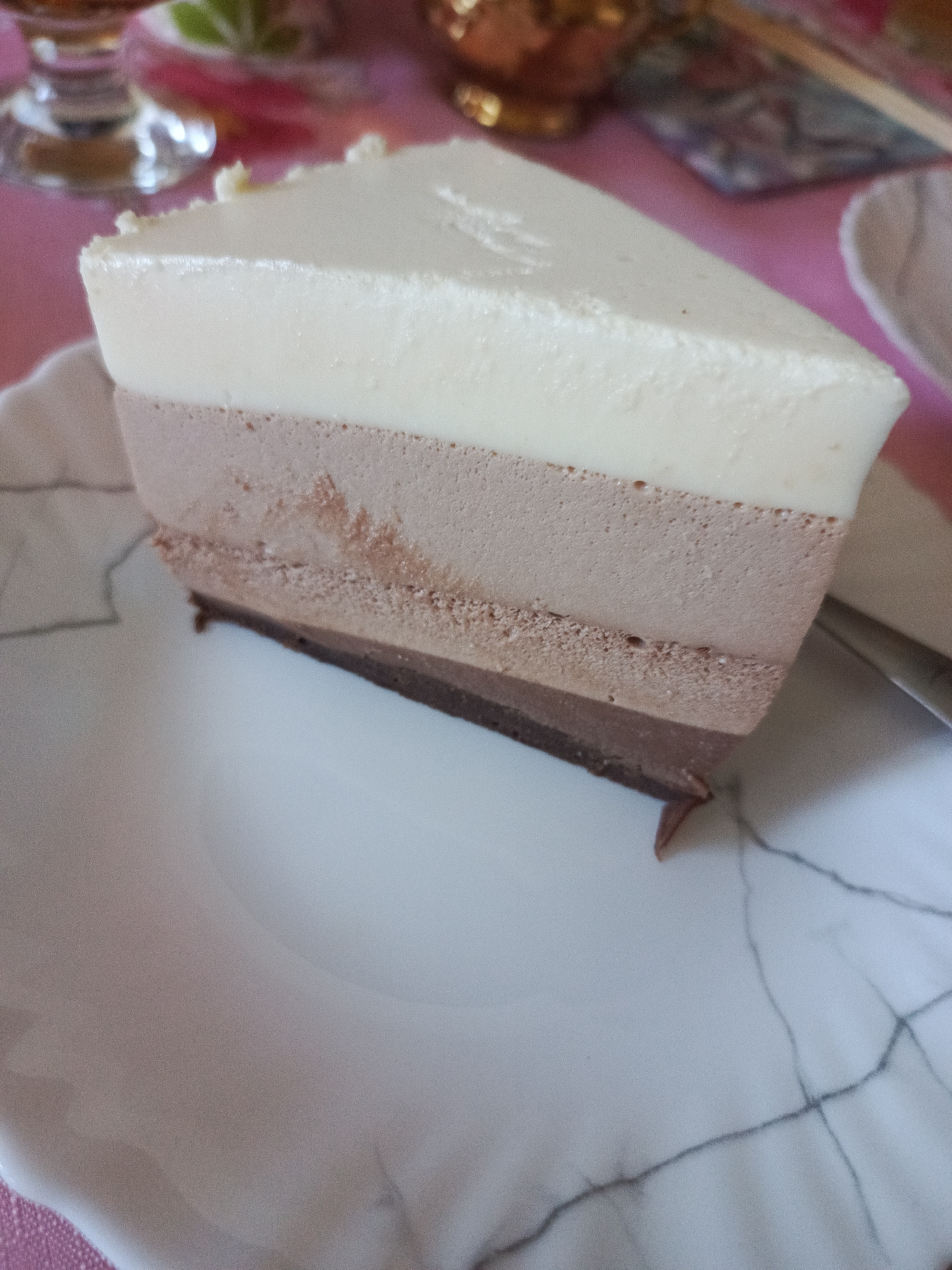 A slice of triple chocolate mousse cake sits on a white plate with a marble-like pattern. The cake has three distinct layers: white on top, milk chocolate in the middle, and dark chocolate at the bottom. The cake is triangular and presented on a pink tablecloth. The background is blurred but suggests a tabletop setting with hints of glassware and decorative items. The scene appears to be indoors.