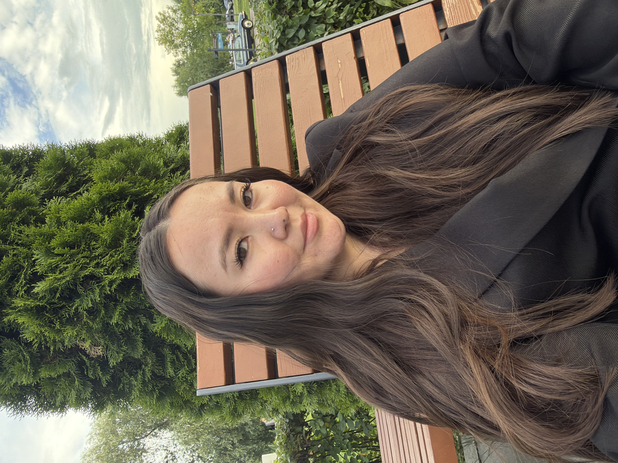 A woman with long, dark brown hair sits on a wooden bench in an outdoor setting. She is wearing a black blazer. Behind her, a large green coniferous tree stands tall, and lush green bushes are visible. In the background, there are other trees, a cloudy sky, and a dark-colored vintage car parked near a streetlamp. The scene appears to be in a park or an area with significant greenery.