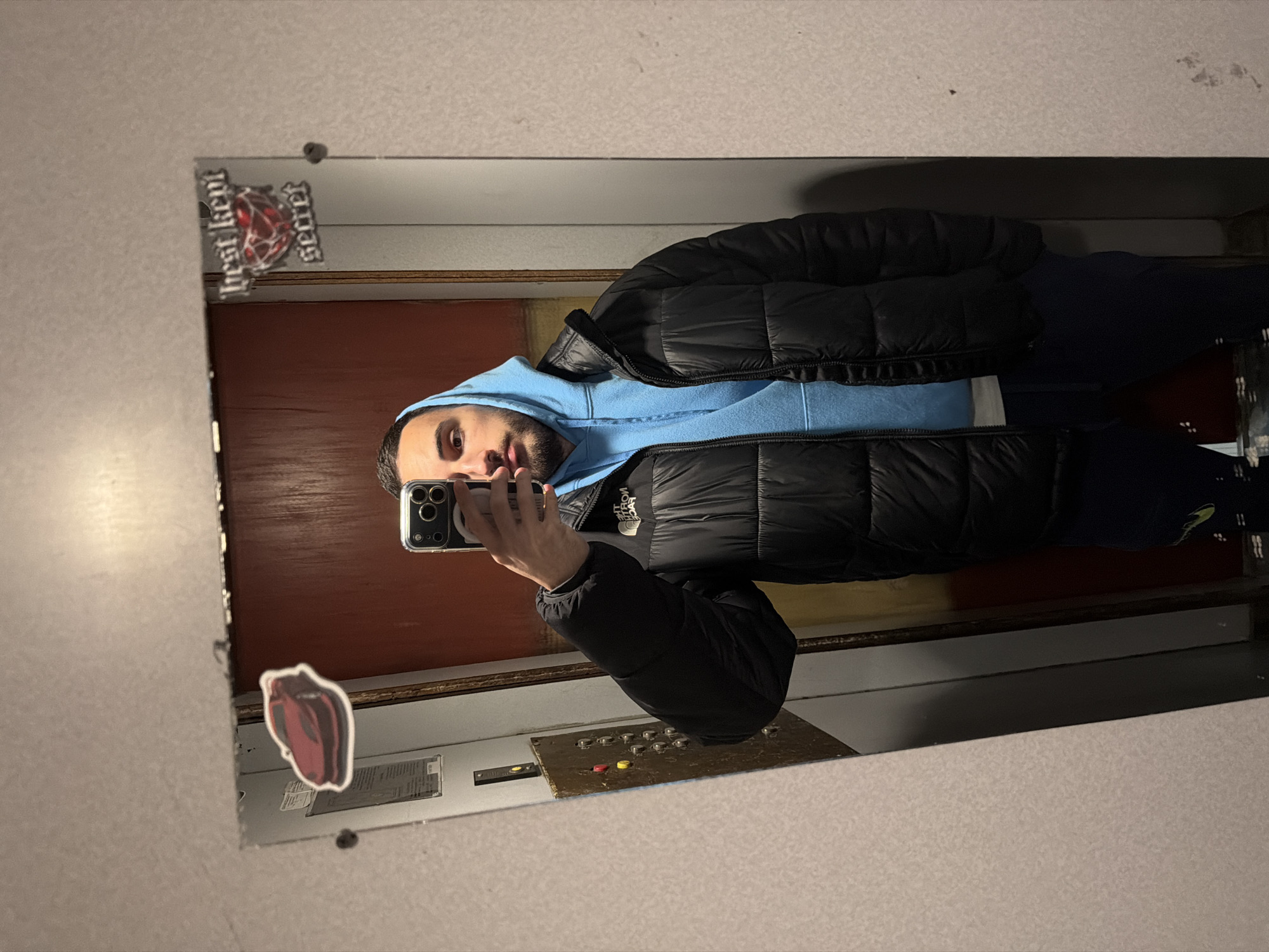A person in a light blue hooded sweatshirt and black puffer jacket is taking a selfie in a mirror. The mirror is located inside an elevator. The elevator has a control panel with buttons. Stickers are affixed to the mirror, including one that reads "best kept secret." The background shows a brown paneled wall within the elevator. The location is Plovdiv, Bulgaria is not indicated in the image.