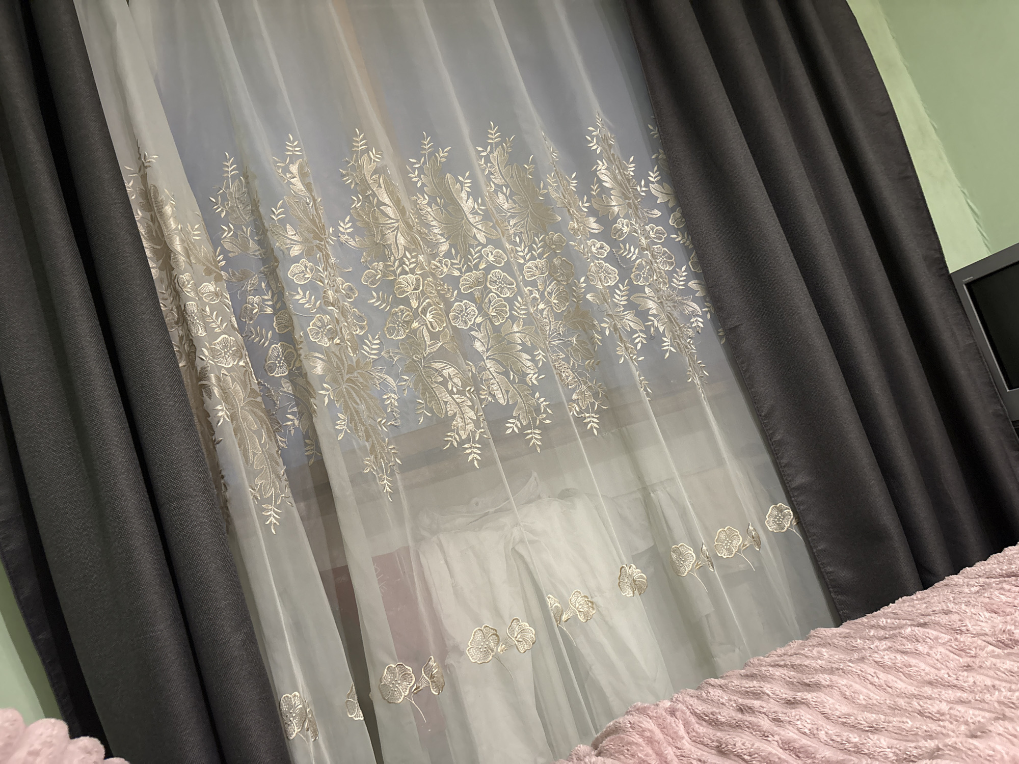 The image depicts a bedroom interior, likely in a residential setting. Two types of curtains adorn a window. The outermost curtains are a dark gray, thick fabric that blocks light. Behind these, a sheer, white curtain with an embroidered floral pattern hangs. The room's walls are painted a pale green. A portion of a television screen is visible against the wall. In the foreground, a pink, textured blanket covers what is probably a bed. While the image suggests a domestic scene, there's no indication of people or specific actions. The image provides no information to verify whether the scene is located in Vsevolozhsk, Russia.