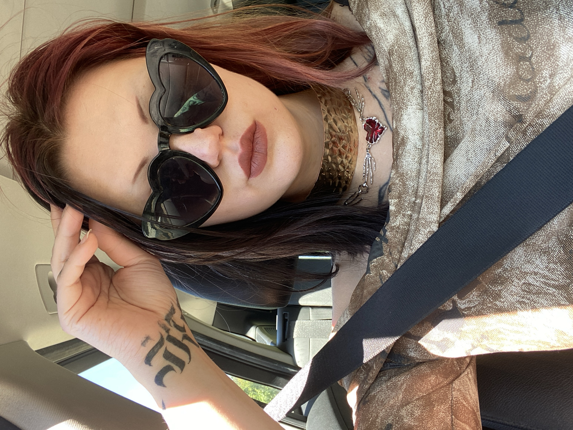A woman with red-tinted brown hair is seated in a car, wearing a black seatbelt and a gold mesh garment. She has heart-shaped sunglasses and a wide gold choker with a red heart pendant hanging below. A tattoo is visible on her left wrist. The interior of the car is visible, with the car door and the external environment seen through the window. The woman is not in Krute Ulcinjske - Krytha e Ulqinit, Montenegro.