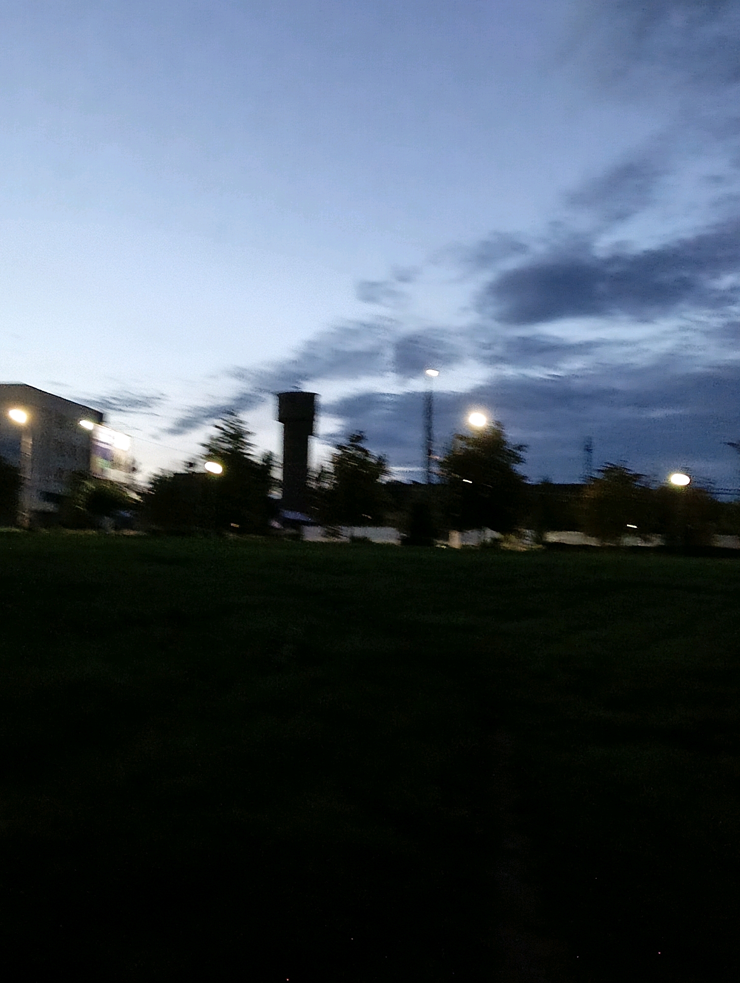 The image depicts an outdoor scene at dusk or dawn, with a dark, grassy foreground. In the middle ground, there is a line of trees silhouetted against the sky. Behind the trees, several man-made structures are visible, including a tall, cylindrical water tower, a building with illuminated windows, and multiple streetlights. The sky is a mix of pale blue and dark, cloudy patches. The overall impression is one of a somewhat industrial or residential area at the edge of a park or open field, possibly in the city of Hrodna, Belarus, given the commonality of such water towers in the region. The image is slightly blurred, suggesting motion during the capture. No people are visible in the frame.