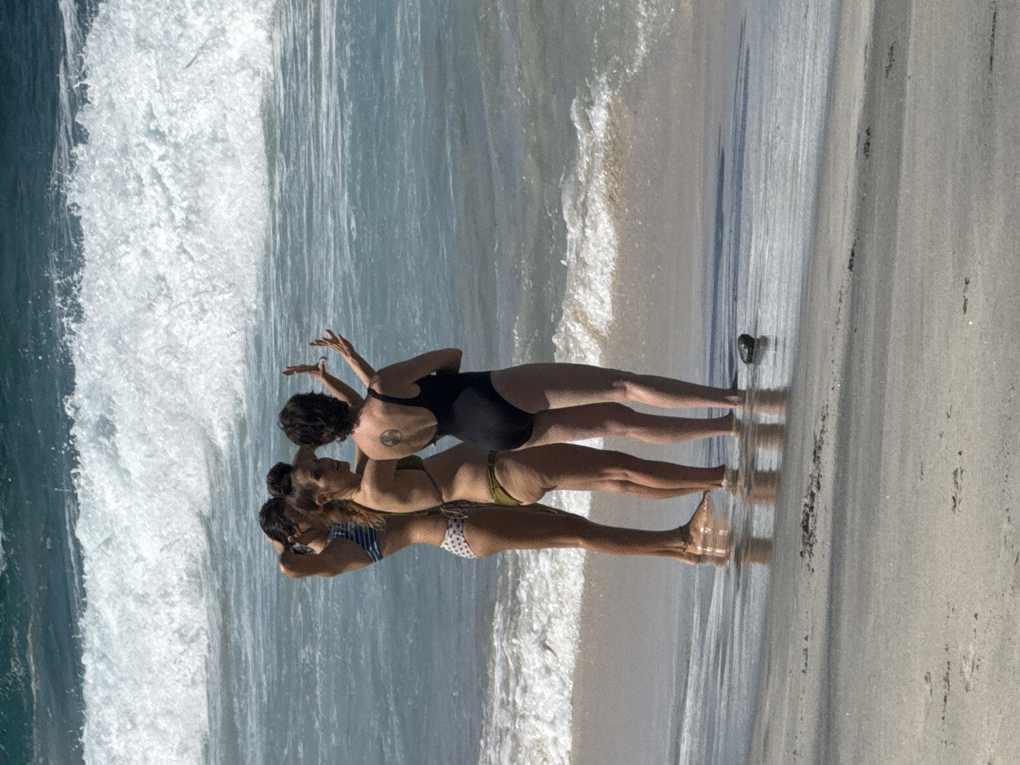 Three women in swimwear stand on a sandy beach, facing the ocean and taking a selfie. The woman on the left wears a blue and white striped bikini. The middle woman is in a green bikini. The woman on the right is in a black one-piece swimsuit. The ocean behind them is a vibrant blue, with white waves crashing on the shore. The sun is shining, creating reflections on the wet sand. There is a dark, pebble-like object on the wet sand to the right of the women's feet.