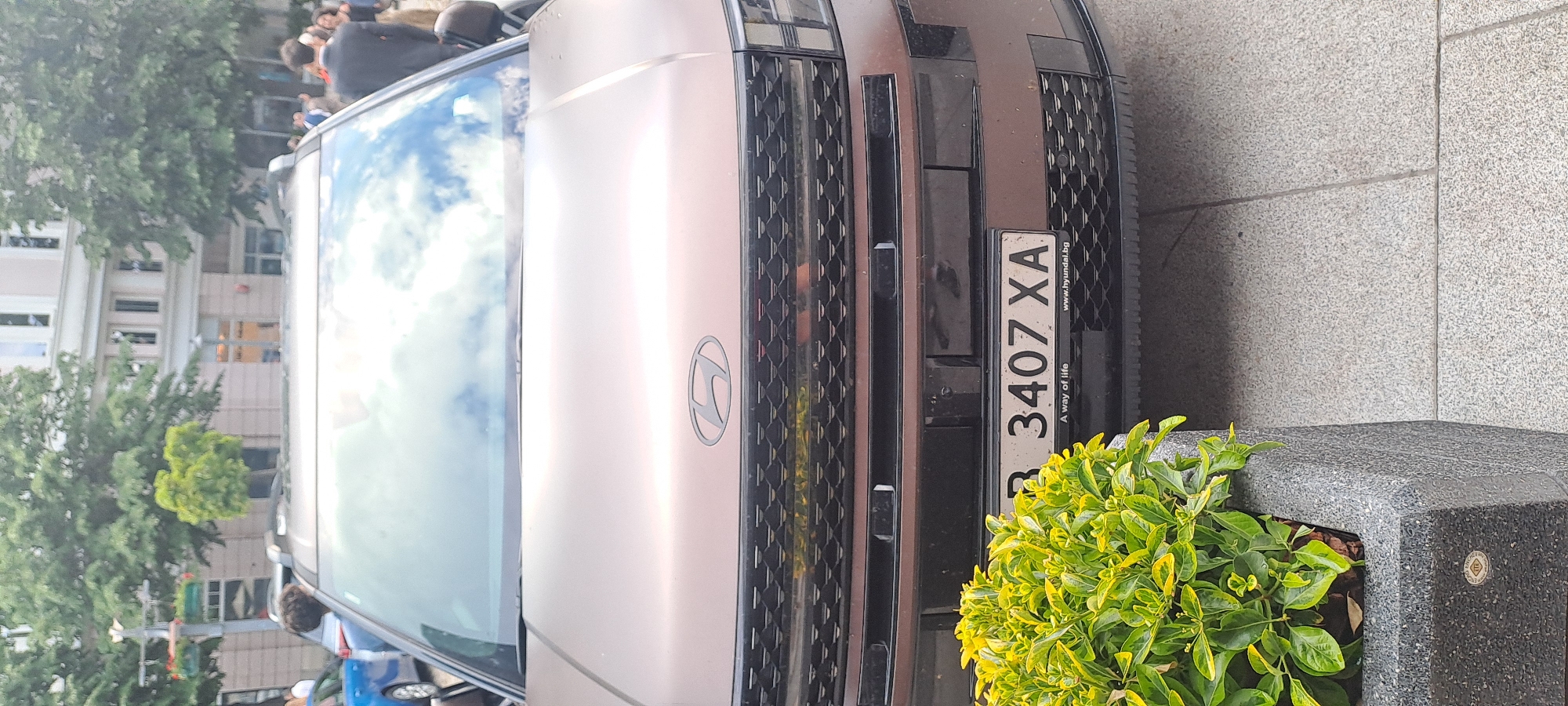 The image shows the front of a light brown Hyundai car with Bulgarian license plate "B 3407 XA". The car is parked on a grey tiled surface, next to a dark grey stone planter with green and yellow leafy plants. Buildings and trees are visible in the background, along with a crowd of people. The license plate indicates the car is registered in Burgas, Bulgaria.