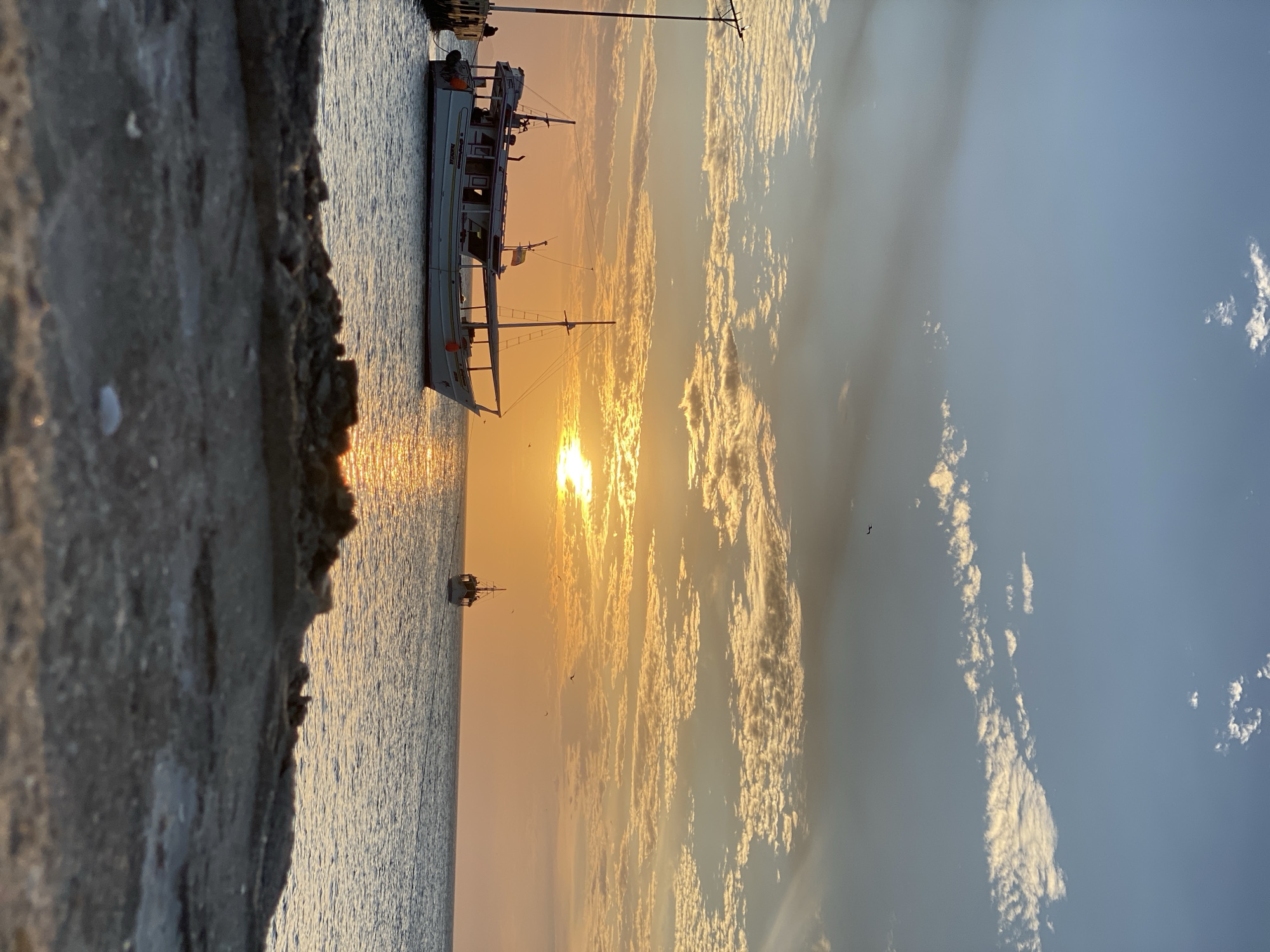 The image captures a sunset scene in Juan Griego, Venezuela. A boat is docked near a concrete barrier. The setting sun is partially obscured by clouds. The water reflects sunlight. There are other boats further out at sea. Several birds are flying in the sky.