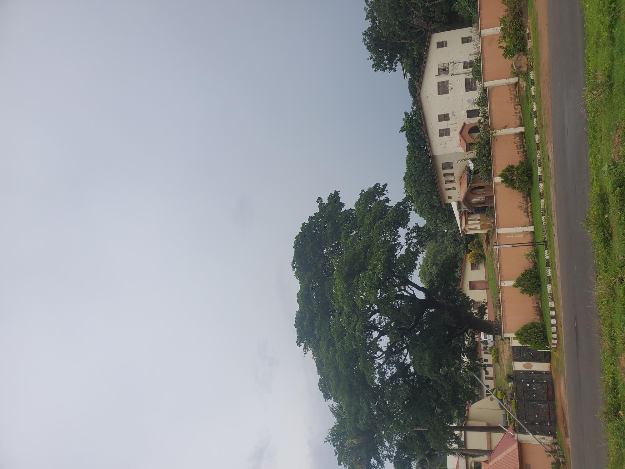 The image depicts a scene in Jos, Nigeria, likely on a cloudy day. In the foreground, a paved road runs horizontally across the bottom of the frame, bordered by green grass and weeds. Behind the road is a light brown wall topped with a decorative white trim, serving as a boundary for a property. Along the wall, neatly trimmed bushes and some taller greenery are visible.
Beyond the wall, several buildings are present. On the right, a large, two-story building with a cream-colored facade and brown-trimmed windows stands prominently. It has a somewhat weathered appearance, with some vegetation growing around its base. To the left of this building, a lower, cream-colored building with brown trim and doorways can be seen, partially obscured by lush green trees. Further to the left, another cream-colored building with red-tiled roofs and dark metal gates is visible, with a large, mature tree with dense green foliage dominating the middle ground of the image. The branches of this tree spread widely, casting shadows on the ground. In the background, more trees and buildings are scattered, suggesting a developed area. The sky above is overcast and a uniform light gray, with no discernible features. A white van is parked in front of one of the buildings in the middle distance. The overall impression is one of a residential or institutional area with established landscaping.