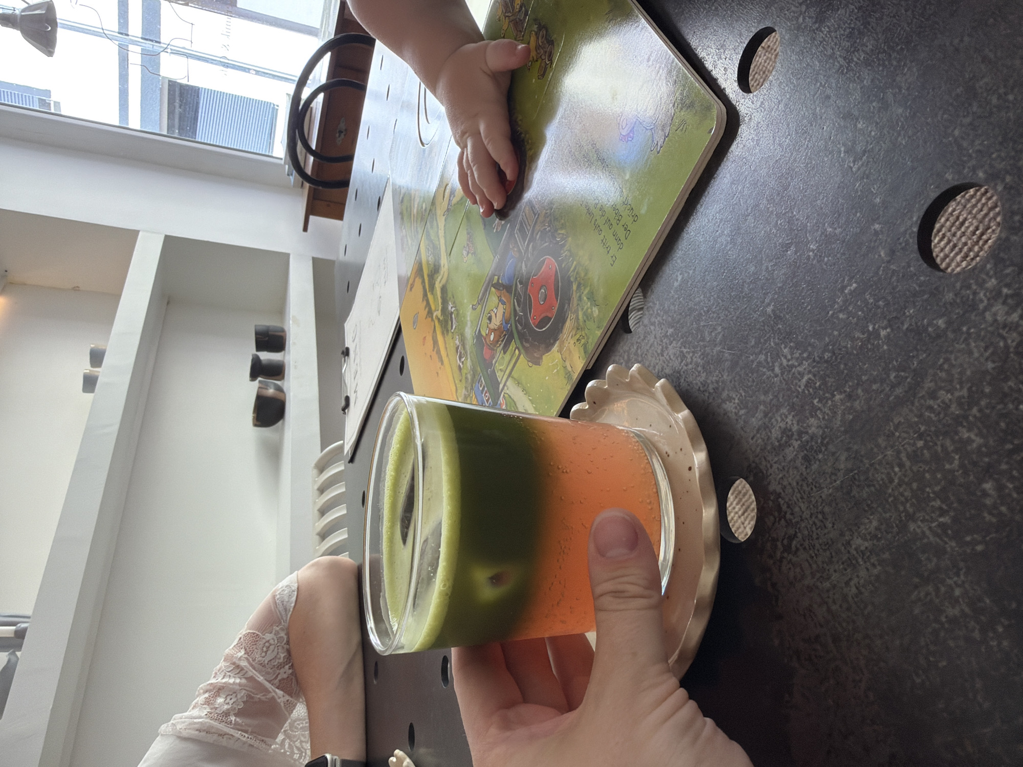 A hand holds a layered drink with green on top and orange on the bottom, with ice cubes, in a clear glass set on a decorative coaster. The drink is on a dark, textured table. To the right of the drink, a children's book with colorful illustrations is open, featuring a tractor and animals. A baby's hand is touching the book. In the background, a person wearing a white top with lace sleeves is visible. Shelves with dark ceramic cups are on the wall. A window with blinds and an external view is also present. The scene appears to be an indoor setting, possibly a cafe or restaurant, in Kaiserslautern, Germany.