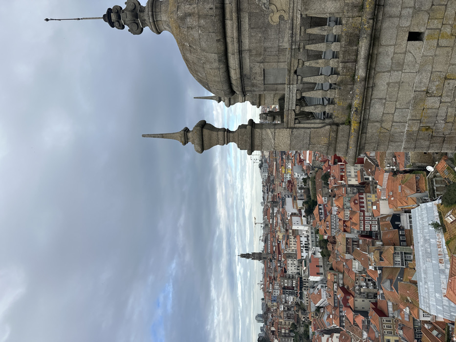 The image displays an elevated panoramic view of the city of Porto, Portugal, captured on an overcast day.
In the immediate foreground, on the right side, a substantial grey stone architectural structure is visible. This includes a rounded dome element topped with an ornate, multi-tiered stone finial and a metal lightning rod or spike. Adjacent to it, a stone balustrade with multiple balusters runs horizontally, and further left, a tall, pointed stone finial rises. The stonework exhibits weathering and some moss growth.
The midground and background present a dense urban sprawl characteristic of Porto. Numerous buildings with a predominance of terracotta-orange tiled roofs stretch across the frame, descending towards the left and center. Buildings feature varied facades, primarily in muted earth tones, white, and shades of red. Streets and alleys are implied by the arrangement of structures.
Dominating the distant city skyline, slightly to the left of the center, is the distinctive tall baroque bell tower of the Clérigos Church (Torre dos Clérigos), confirming the location as Porto. Several construction cranes are visible in the further distance, indicating ongoing development.
The sky is largely overcast with extensive grey cloud cover, though traces of lighter blue sky are faintly visible towards the distant horizon on the left. No individuals or specific actions are discernible within the frame.