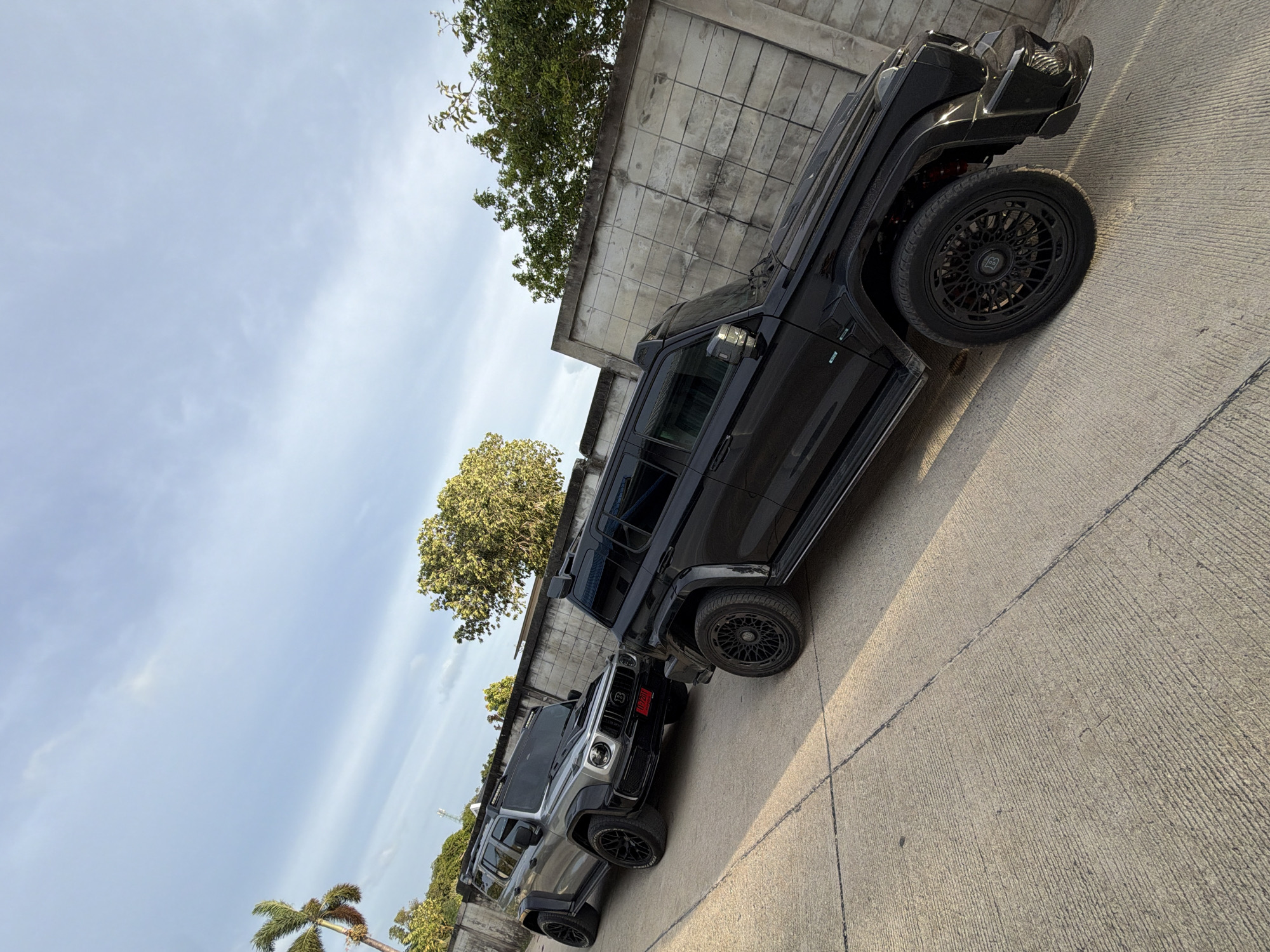 The image depicts two heavily modified Mercedes-Benz G-Class vehicles parked on a concrete surface in a city area. The vehicle in the foreground is black and features an aggressive body kit, custom black wheels with a intricate spoke design, and a prominent roof rack. The second vehicle, partially obscured by the first, is silver with similar custom black wheels and also appears to have a roof rack. Both vehicles have a rugged, off-road aesthetic.
Behind the vehicles is a textured concrete wall with a grid pattern. Trees with lush green foliage are visible above the wall, and a palm tree is present in the background on the left. The sky is a pale blue with some wispy clouds, suggesting it is either early morning or late afternoon. The lighting indicates sunlight casting shadows on the ground. The scene suggests an outdoor parking area, possibly in a residential or commercial district. Given the context of Ko Samui District, Thailand, this could be a private property or a designated parking space in a tropical urban setting. There are no people visible in the image.