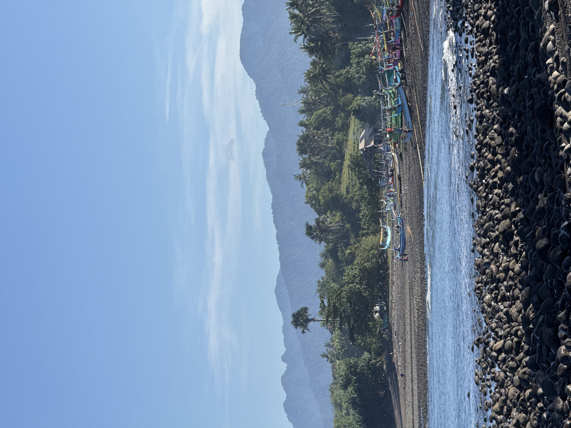 The image depicts a coastal scene with a rocky beach in the foreground. The beach is composed of dark, rounded stones. A narrow strip of water is visible between the stones and the land. Beyond the beach, there is a line of vegetation, primarily consisting of trees and shrubs. Along the edge of the vegetation, there are numerous fishing boats. In the background, there are layered mountains visible under a blue sky. The location appears to be a remote, natural setting with signs of human activity in the form of the boats.