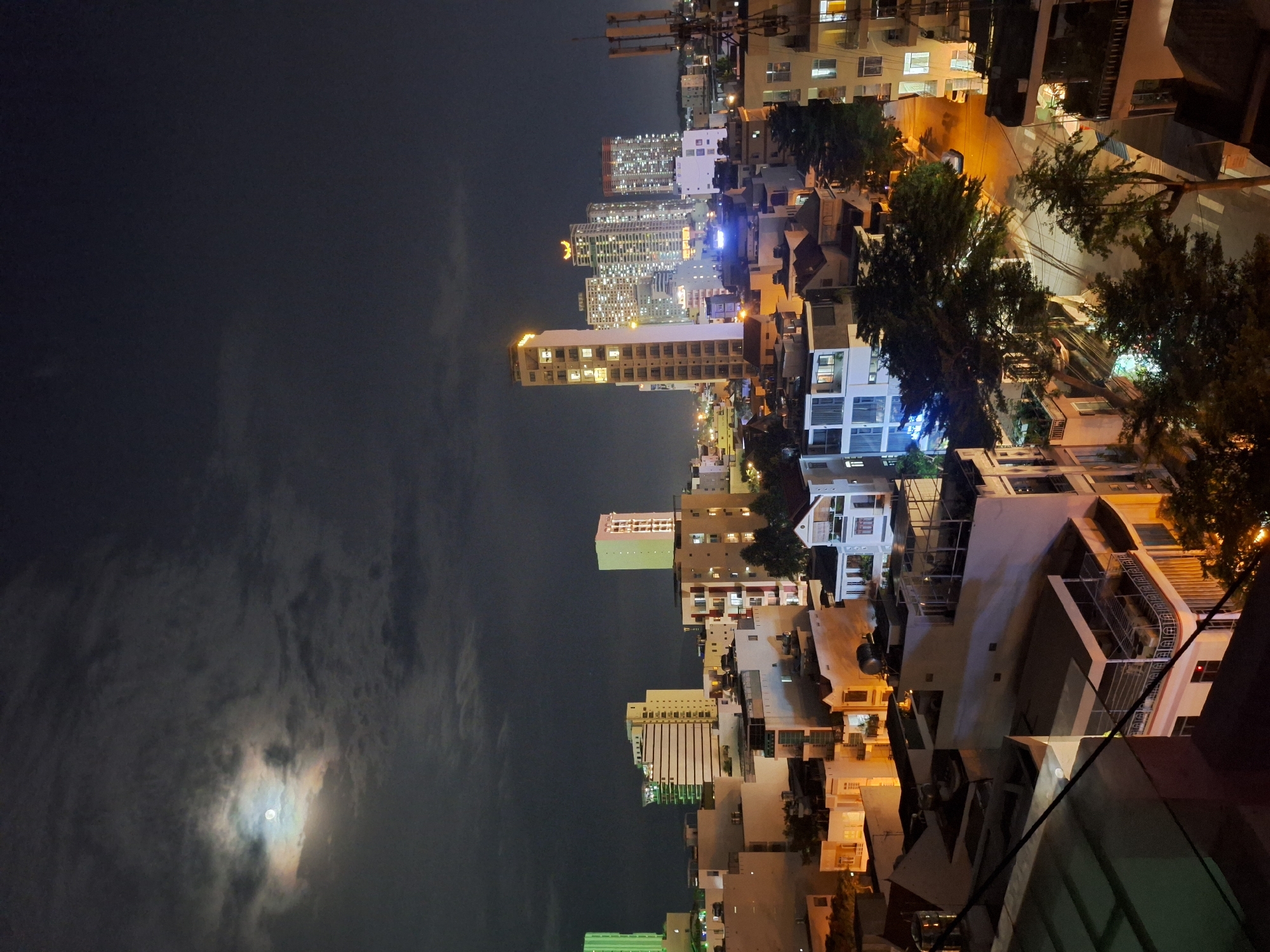 This is a nighttime, high-angle, long-exposure photograph of the city of Nha Trang, Vietnam, taken from a rooftop. A full moon is visible in the upper left quadrant of the sky, partially obscured by clouds. The city skyline is illuminated by numerous lights from buildings and streetlights. The foreground shows rooftops of residential buildings and a street lined with trees. In the middle ground, there are several mid-rise and high-rise buildings, some with illuminated windows and facades. The background is dominated by the dark sky and more distant city lights, suggesting a densely populated urban area. The overall impression is one of a vibrant city at night under a full moon.