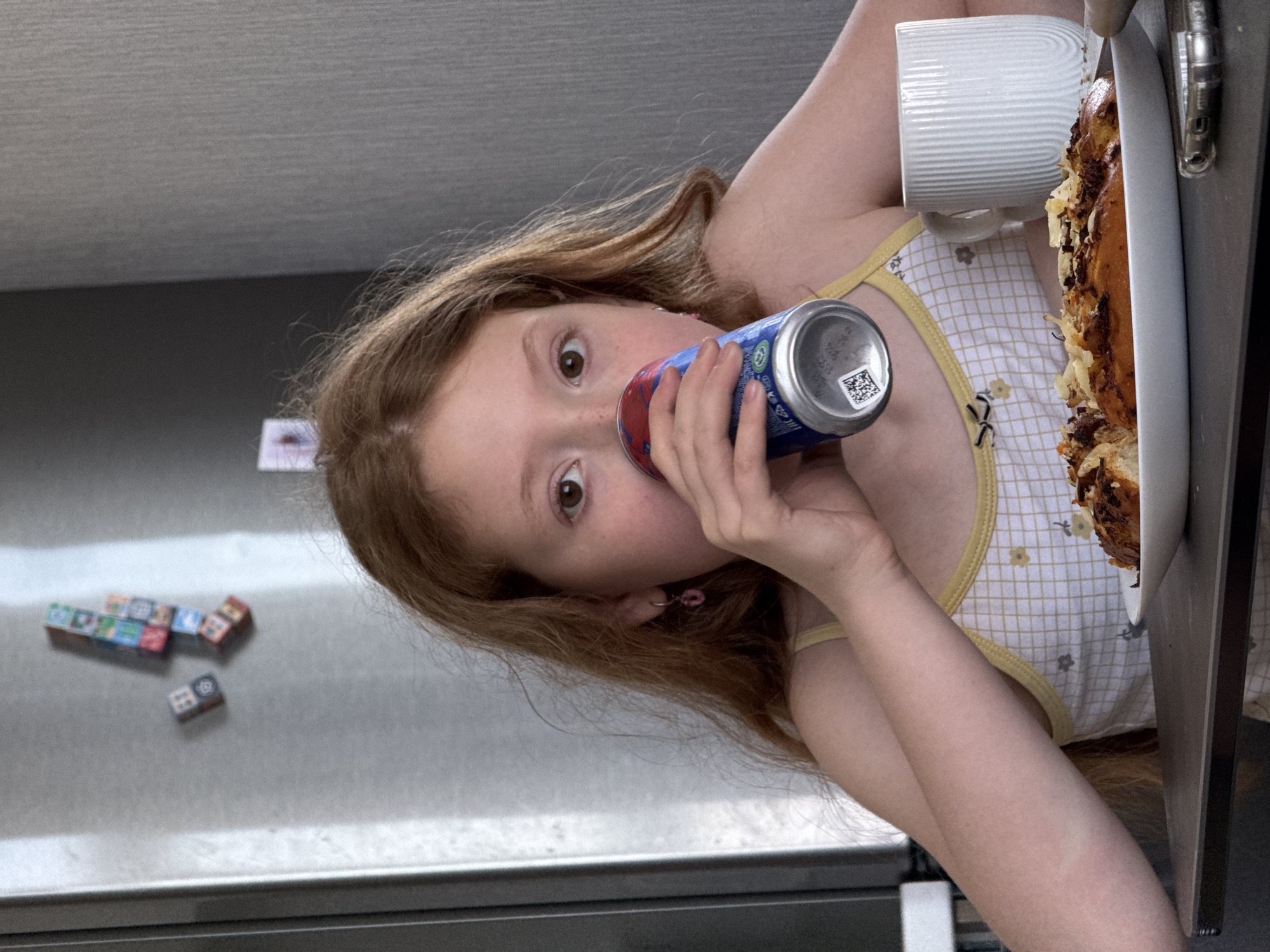 A young, red-haired girl is positioned in the foreground, her face slightly turned to the side as she holds a blue and red can up to her mouth. Her eyes are wide open and directed toward the viewer. She wears a white and yellow checkered top with small floral patterns. To her right is a white mug and a plate with what appears to be a pastry or baked dish on it. Behind her, a silver refrigerator is visible, adorned with several colorful square magnets and at least one small die. The location is presumed to be an indoor setting. The location of Rostov-on-Don, Russia cannot be confirmed from this image.