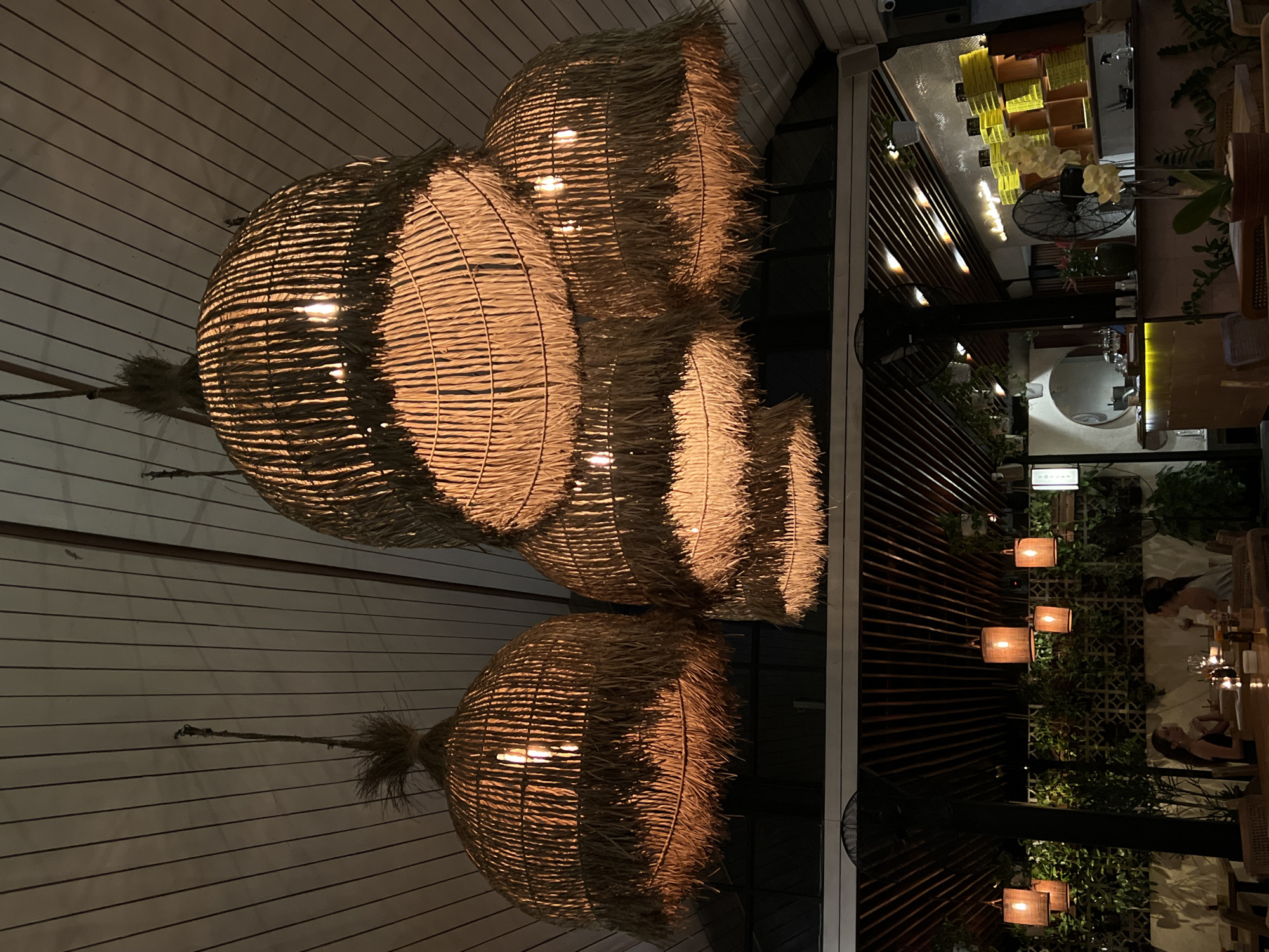 A dimly lit interior view of an establishment in North Kuta, Indonesia, features distinctive lighting fixtures. Four woven pendant lamps, shaped like inverted teardrops, hang from a white plank ceiling, each emitting a warm glow. The lamps are arranged vertically with the largest above and the smallest below. The interior transitions from left to right, moving from the ceiling and light fixtures to a darker dining area filled with greenery. The dining area is partially lit with square lamps along a vertically designed wall. Tables are visible, with people seated, though details are obscured by the darkness. A fan and various plants add to the ambiance. The location is identified as North Kuta, Indonesia.
