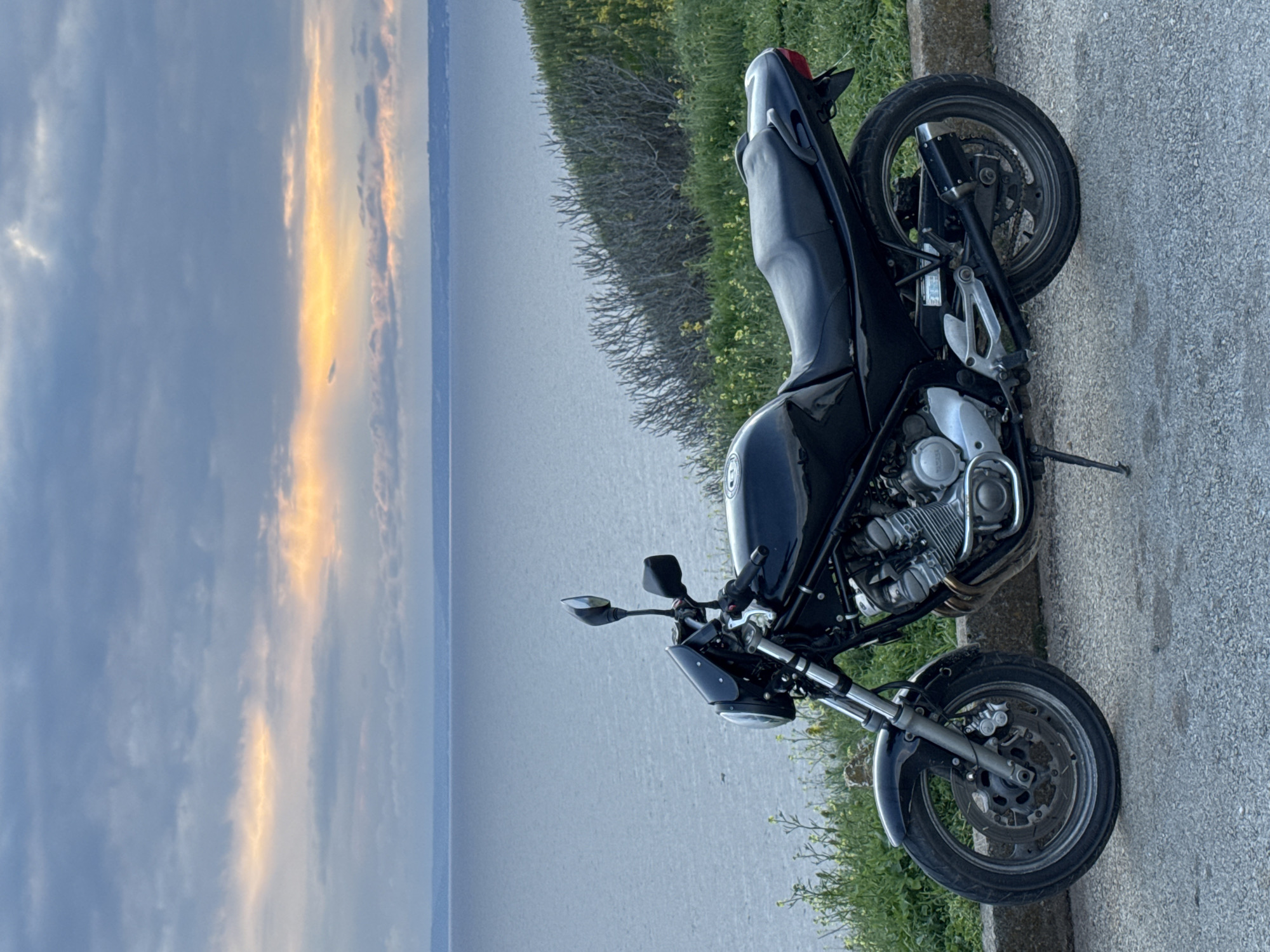 A black motorcycle is parked on the side of a paved road next to a grassy embankment, overlooking a body of water under a cloudy sky with a sunset visible in the distance. The location appears to be Balgarevo, Bulgaria, based on the landscape and vegetation. No people are visible.