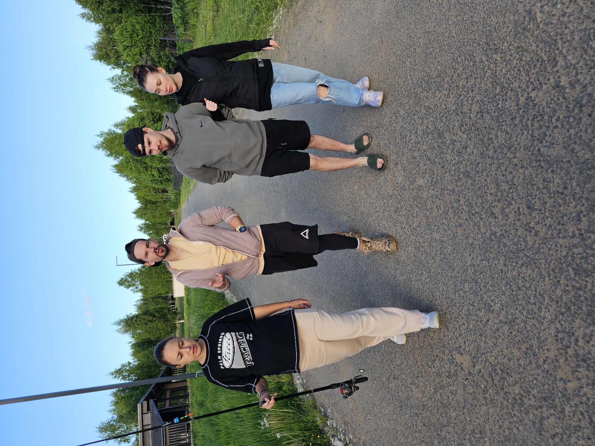 Four people are standing on a paved road. To the left is grass and trees, and a small building. The person closest to the camera is holding a fishing pole. The person at the top of the image is wearing jeans with a hole in them and a black jacket. The location is Мышкино, Russia.