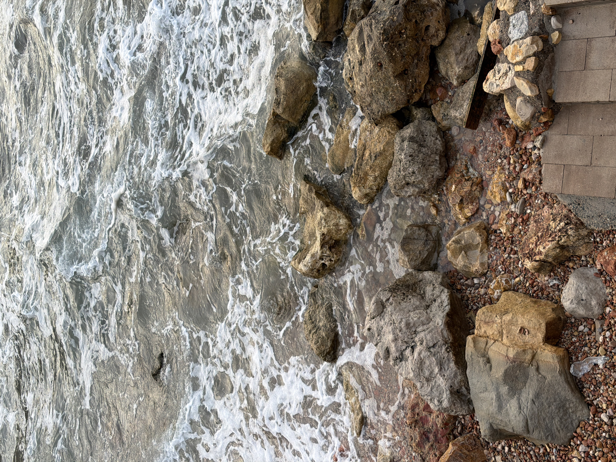 The image depicts a rocky shoreline with waves gently lapping against the rocks. The water is a murky gray-green, and small white foam patches are visible where the waves break. The rocks are of various sizes and colors, ranging from dark gray to reddish-brown and yellow-brown. The ground leading away from the water is covered with smaller pebbles and stones. In the lower right corner, there are a few concrete steps, suggesting a constructed pathway or access point. There are no people visible in the image. The scene appears to be a natural coastal area, possibly within Salou, Spain, given the presence of a Mediterranean-style shoreline with mixed rocky and pebbly terrain.