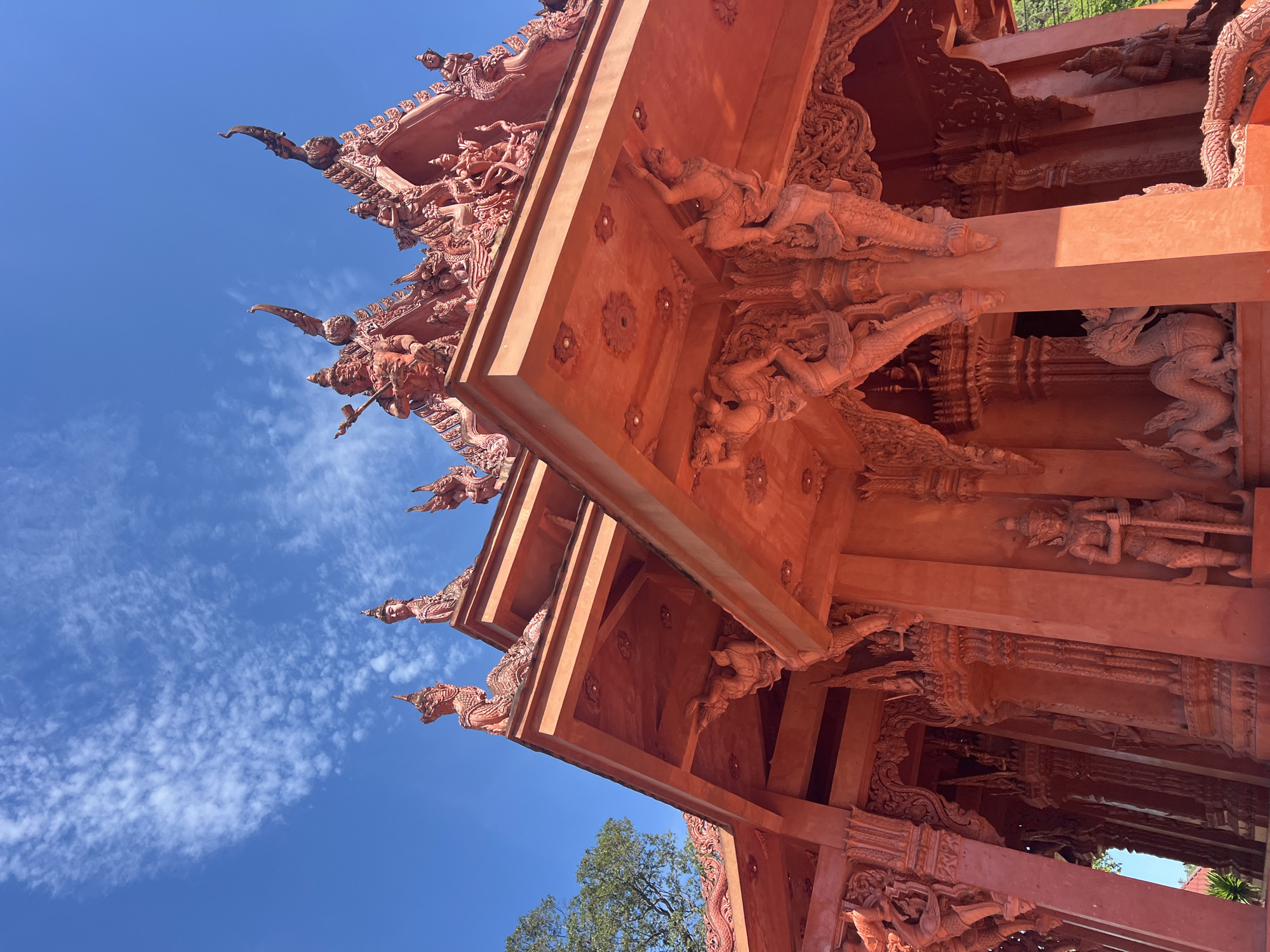 A low-angle, full shot captures the vibrant red exterior of a temple in Baan Hua Thanon, Thailand, under a bright blue sky dotted with wispy white clouds. The temple's ornate architecture is a focal point, with intricate carvings and statues adorning its rooflines, pillars, and eaves. Sculptures of mythical creatures, including serpents and what appear to be deific figures or guardians, are prominently featured. The building exhibits a traditional Southeast Asian design, characterized by its layered roofs and decorative elements. Sunlight casts shadows on the structure, highlighting the textures and details of the sculptures. In the background, a portion of a green tree is visible on the left, contrasting with the warm tones of the temple and the cool blue of the sky. The image is taken from a ground-level perspective, looking upwards at the temple's facade.