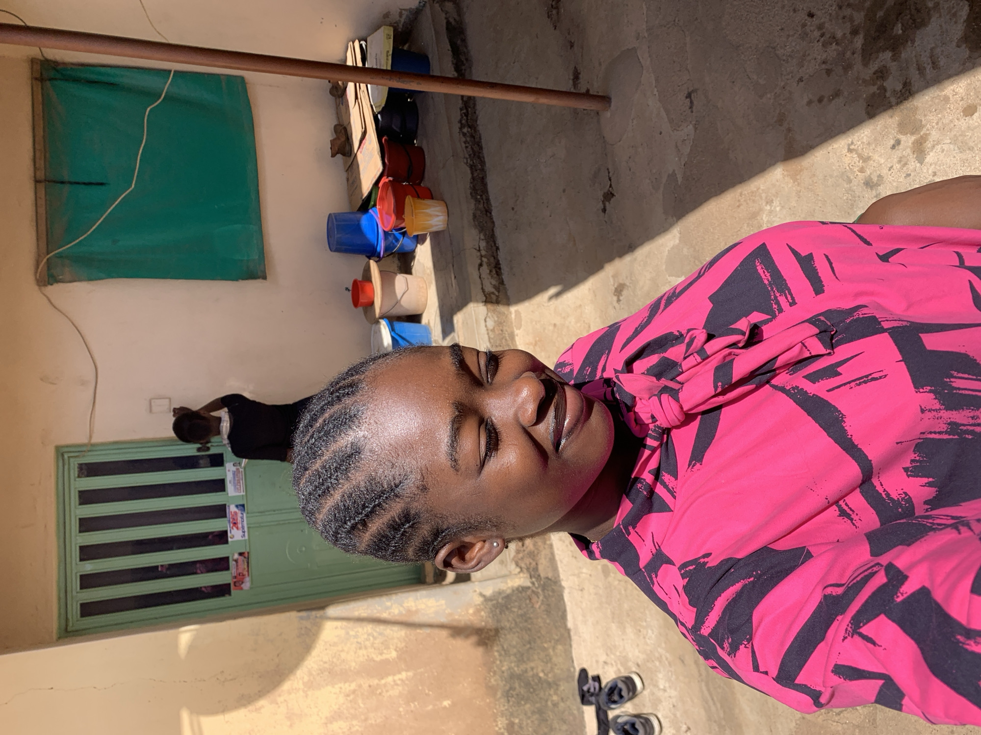A medium shot depicts a woman in a pink shirt with black patterns sitting outside in a sunlit area in Rafin Par, Nigeria. She has her eyes closed and her hair is braided back from her face. Behind her, a green door with barred windows is visible. Next to the door, another person is present. A green fabric is hanging on the wall. A stack of containers is stacked on the ground beside the wall. The ground is made of concrete with a portion covered in shadow. A pair of shoes is on the ground.