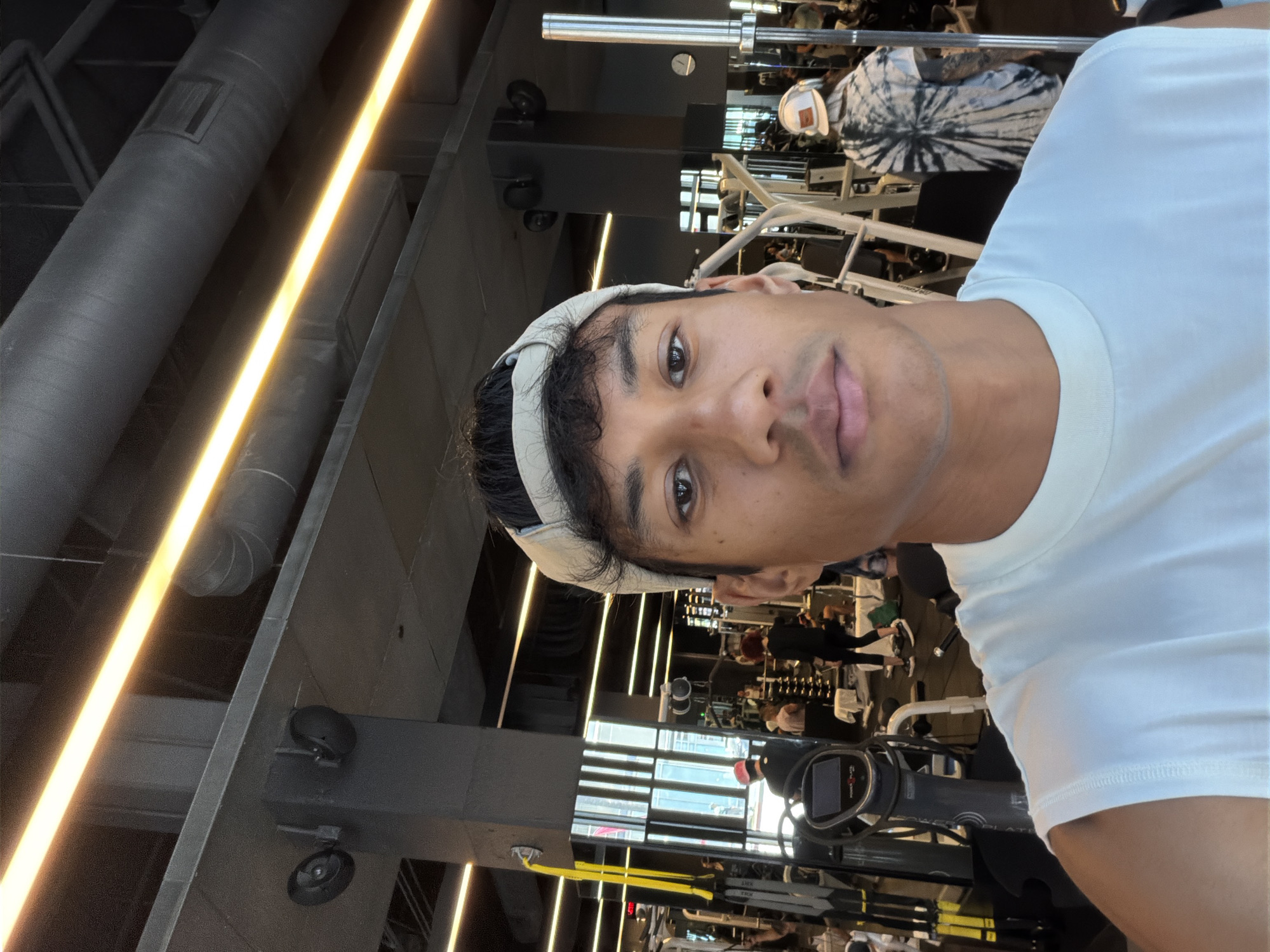 A selfie photo shows a person indoors at a gym in Los Angeles, United States. The person has brown skin, dark hair, and is wearing a white headband and a white T-shirt. The gym contains exercise equipment, including weight machines and treadmills. Overhead lighting illuminates the scene. Other individuals are present in the background.