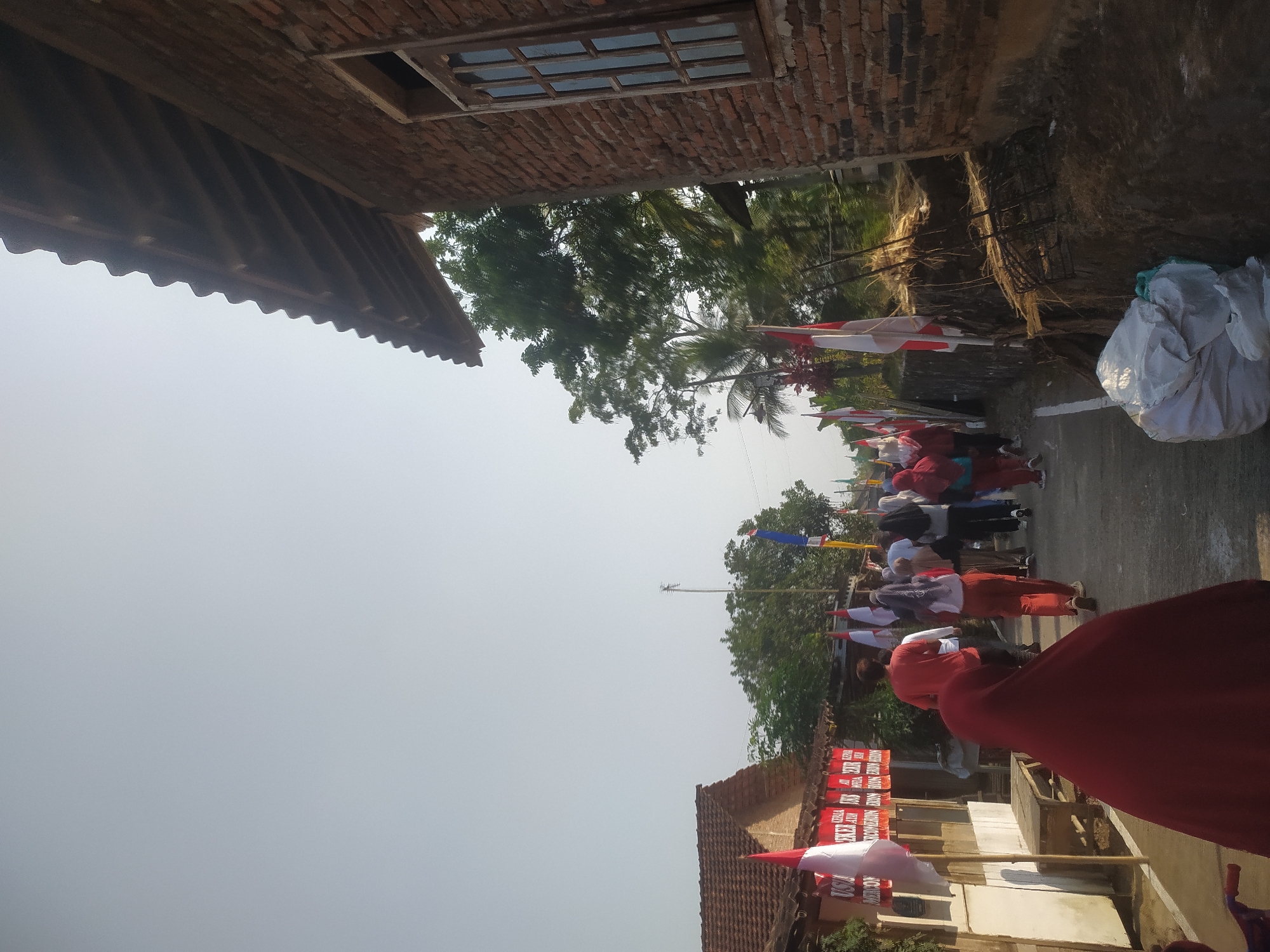 The image depicts a street scene in Lopait, Indonesia, likely during a festive or commemorative event. A group of people, including men and women, are walking along a paved road. Some individuals are wearing red, white, and other colored clothing. Several Indonesian flags, characterized by their red and white colors, are visible, some on poles and others as part of banners displayed on buildings.
On the left side of the road, there are low-lying buildings with tiled roofs, one of which has a white facade with red banners displaying text in Indonesian. A wooden bench is positioned against the building. On the right side of the road, a brick building with a wooden-framed window dominates the foreground. Adjacent to this building, there is a raised embankment covered with dry vegetation and some debris. Further down the road, more trees and foliage are visible, suggesting a semi-rural or suburban environment. The sky is clear and bright, indicating daylight. In the immediate foreground on the right, a large white plastic bag is placed on the ground.