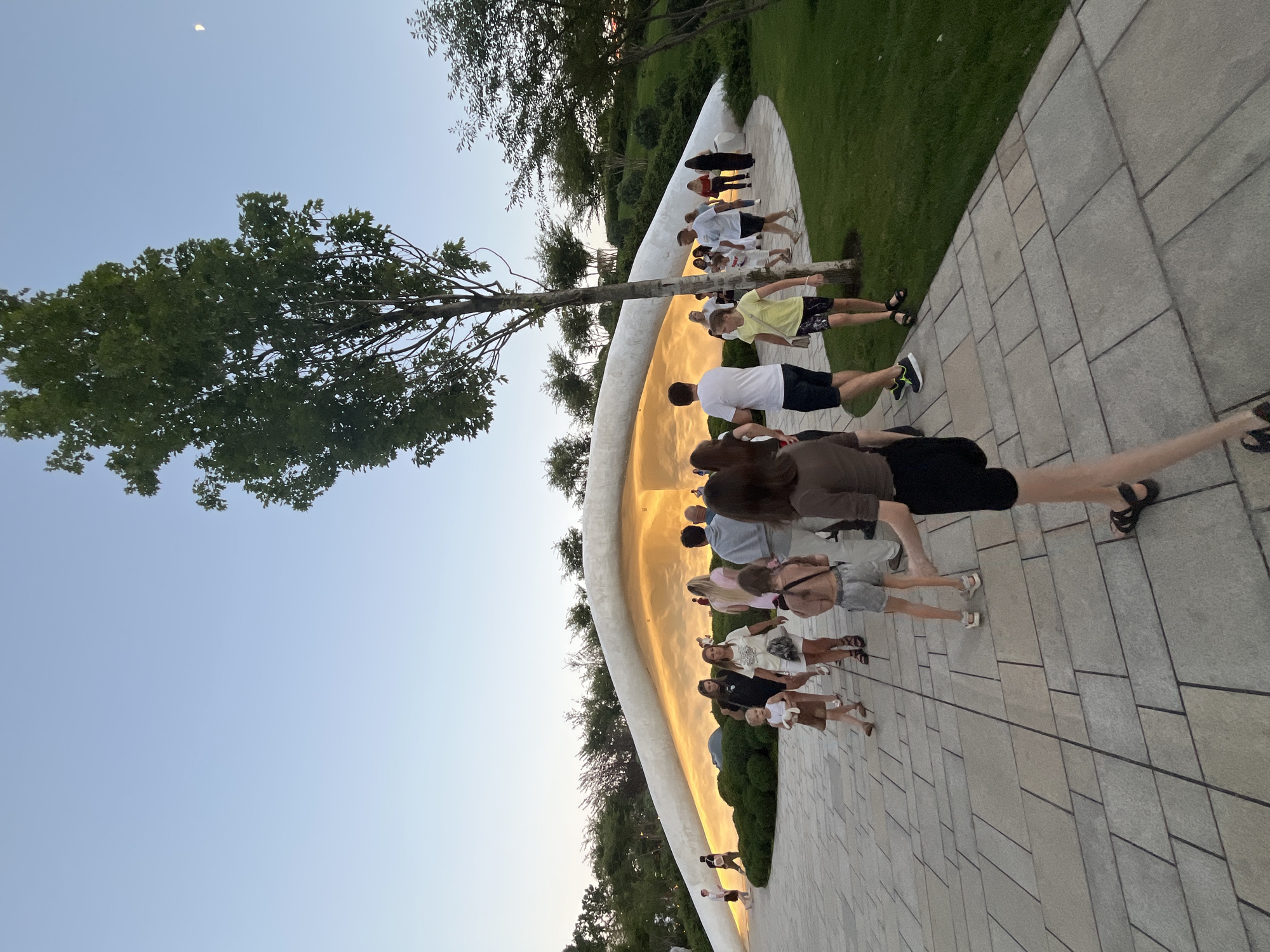 The image depicts a group of people walking along a paved pathway in an outdoor park-like setting. In the background, a large, modern, white structure with an illuminated interior is visible, possibly an architectural feature or building. The sky is clear and shows a crescent moon. Trees and manicured green spaces surround the pathway and the structure. The lighting suggests it is either early morning or late evening. The scene is likely located in the city of Krasnodar, Russia, given the context. The people are dressed in casual summer attire. Some are walking in pairs or small groups, engaged in leisurely activity.