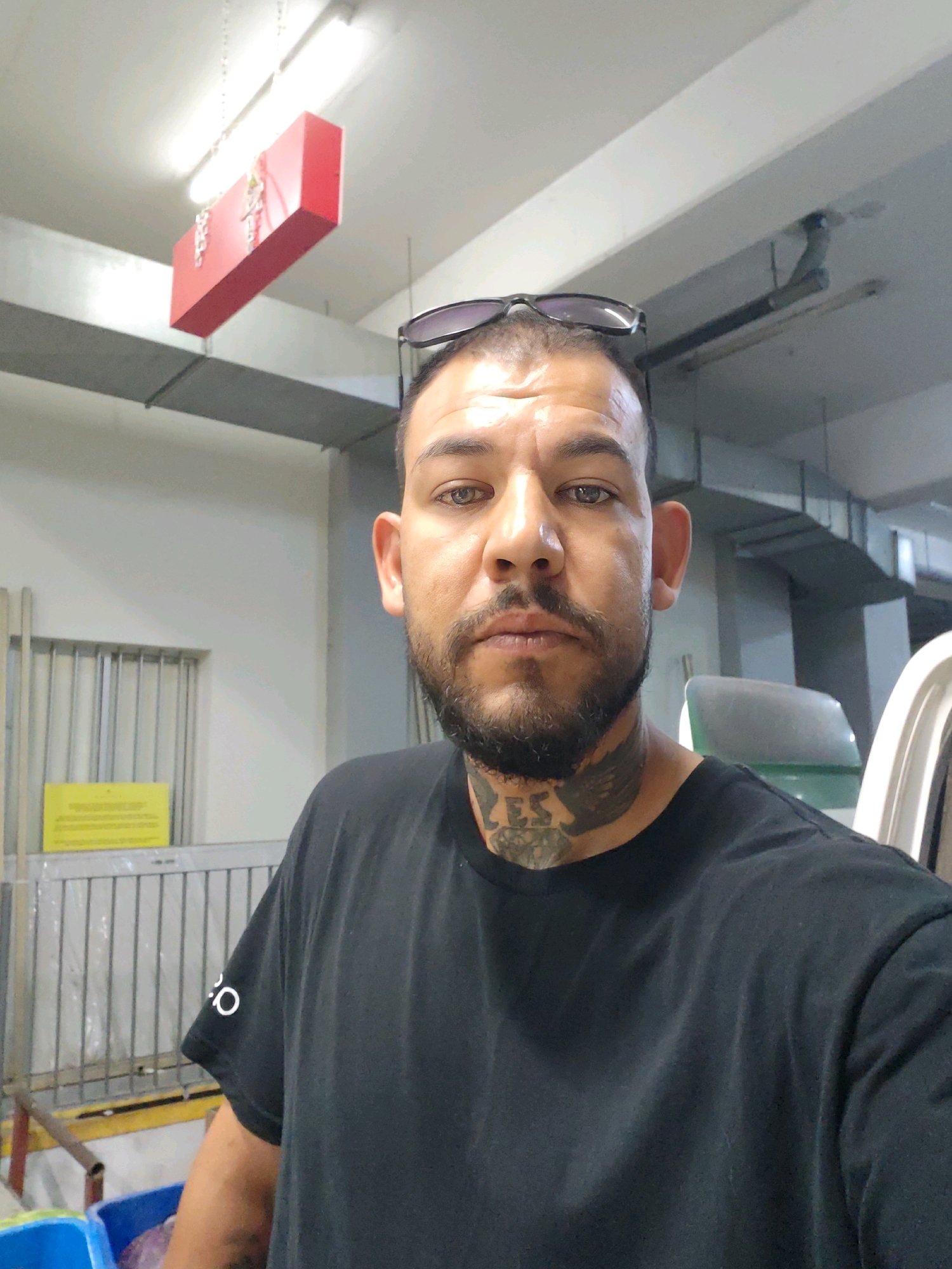 A man with tattoos on his neck and a beard is looking at the camera. He is wearing a black t-shirt and sunglasses on his head. In the background, there is a red box hanging from the ceiling, metal bars, and a yellow sign. The environment appears to be an indoor space, possibly a warehouse or storage area.