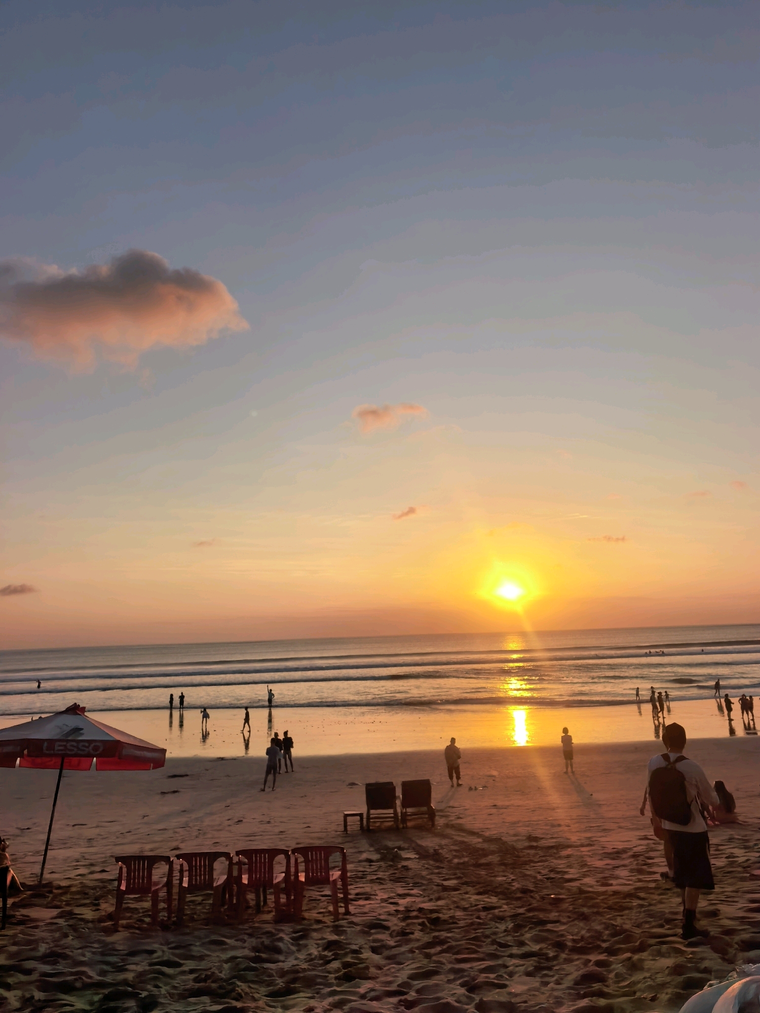 This is a photograph of Kuta Beach in Kuta, Indonesia, during sunset. The sun is low on the horizon, casting a warm orange glow across the sky and reflecting on the water. Several people are visible on the beach and in the shallow water, some walking, some standing, and a few appear to be playing in the waves. In the foreground, there are several red plastic chairs arranged on the sand, and a red and white beach umbrella with the word "LESSO" is set up. The sand is textured and appears damp in some areas. The ocean has gentle waves, and a reflection of the sunset is visible on the wet sand near the water's edge. The sky is a gradient of blue and orange, with a few scattered clouds.
