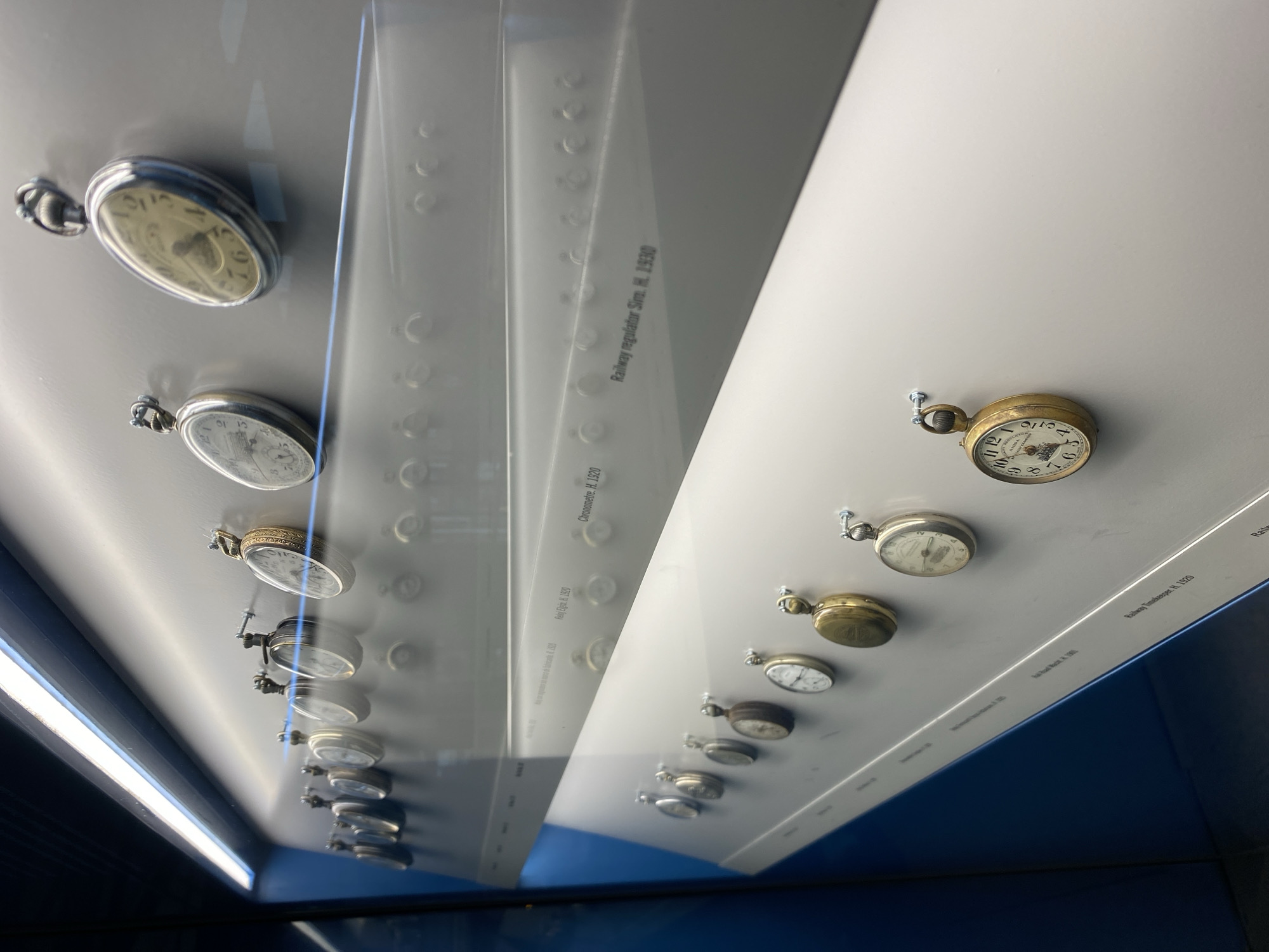 The image shows a display of vintage pocket watches in a glass case. The watches are arranged in a line on a white surface with labels next to them. The labels identify the watches as "Railway regulator Siro. H. 1930", "Chronometre. H. 1920", and "Railway Timekeeper. H. 1920". The location is possibly a museum or exhibit in the city of Gijón/Xixón, Spain. The environment is indoors with blue walls.